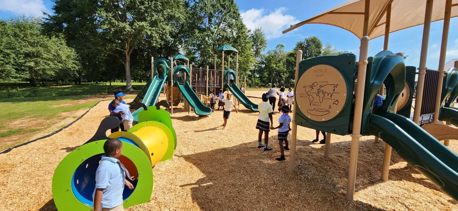 Children playing on a large playground in a park with slides, tunnels, and a world map sign.