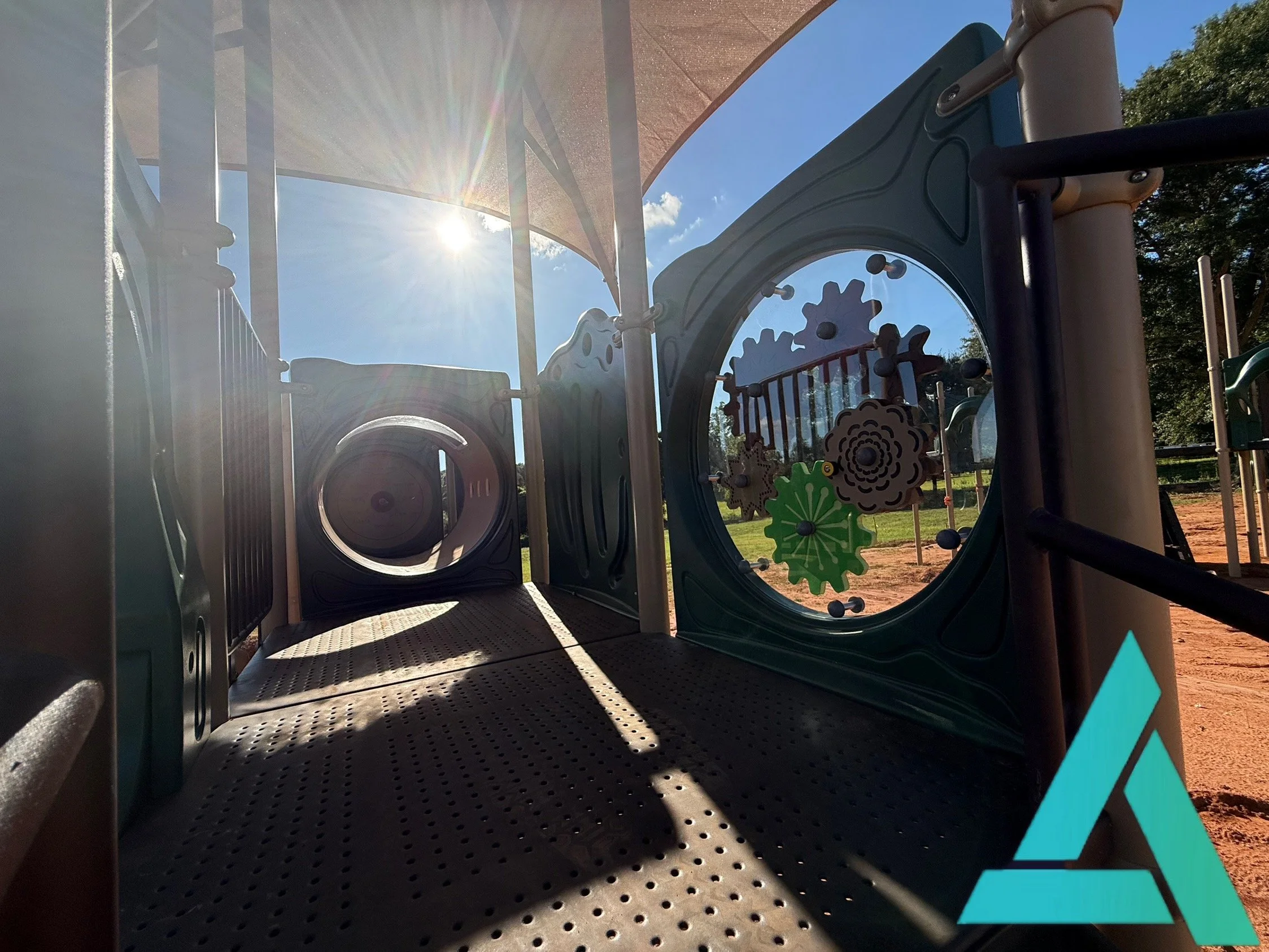 Playground equipment with a tunnel and gear-shaped decorations, under a shade structure on a sunny day.
