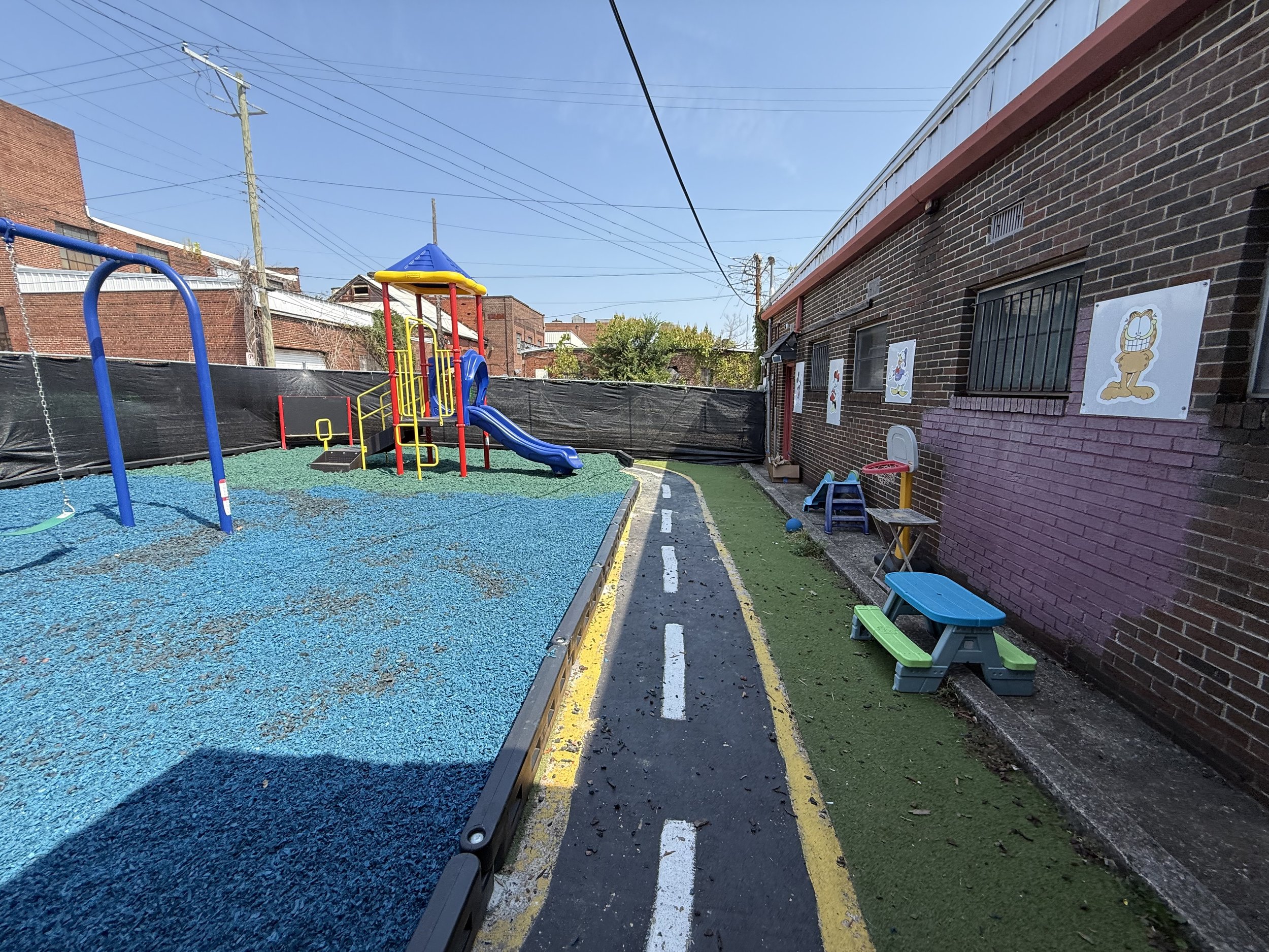 An outdoor playground area with a blue rubberized floor, a colorful play structure with a slide, swings, and a small basketball hoop, enclosed by a black privacy fence. A brick building with cartoon character posters is adjacent to the playground, wi