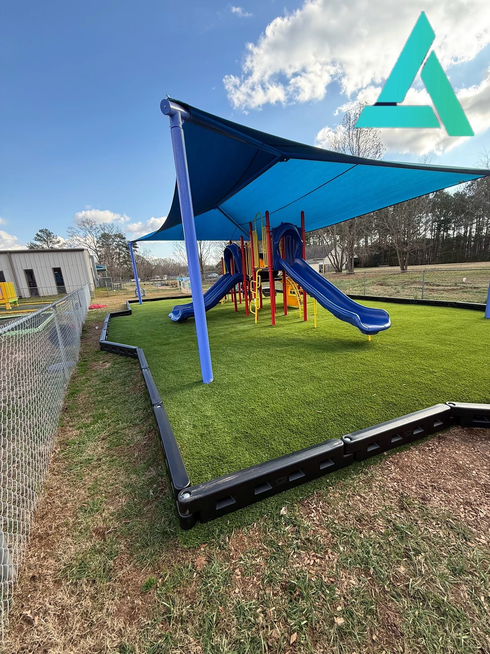 A playground with two blue slides and a blue shade structure, surrounded by a black fence and situated on artificial green turf, with trees and a building in the background, under a partly cloudy sky.
