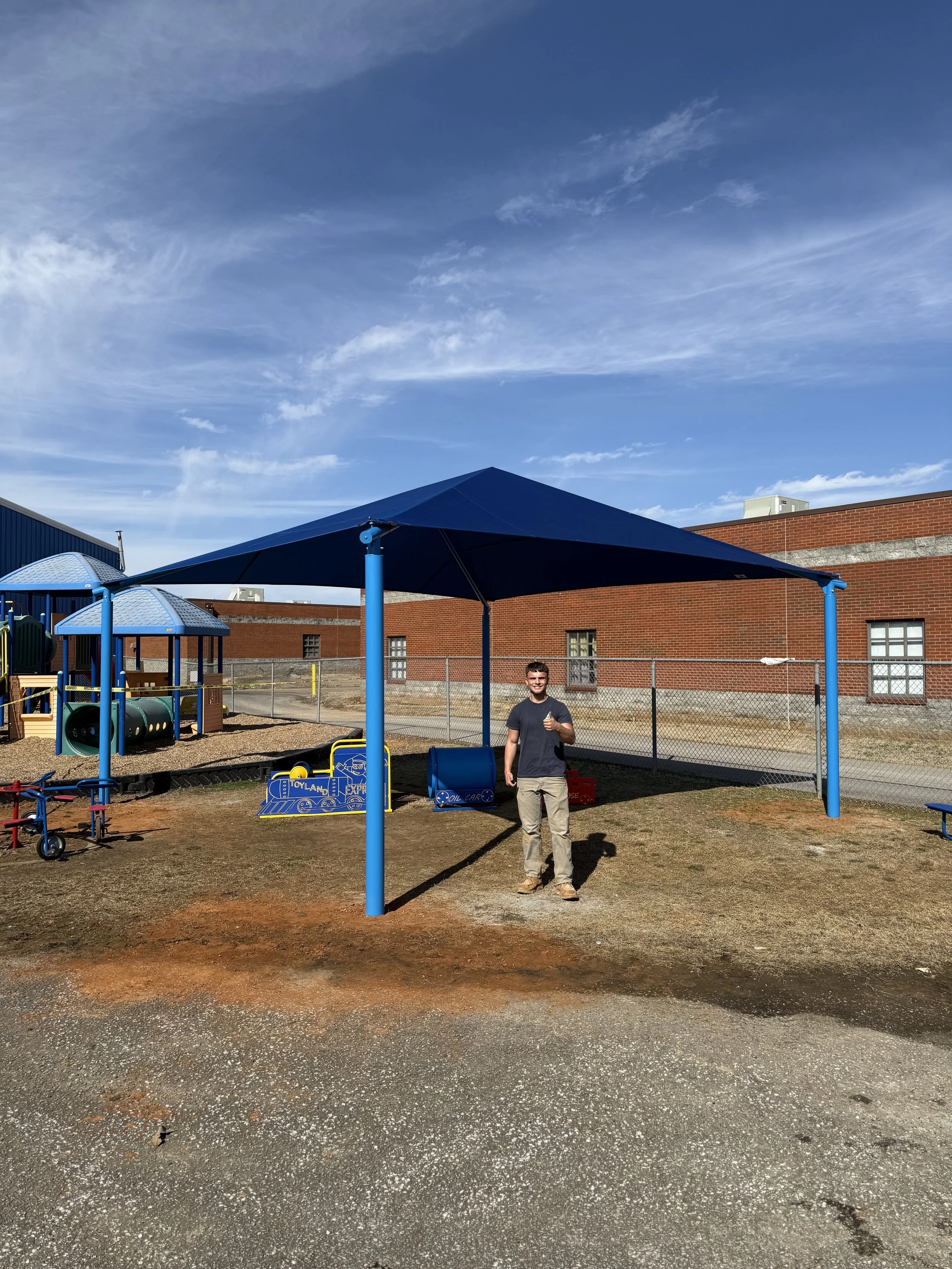 A man holding a drink standing in front of a playground with a large blue canopy, blue poles, and various play structures near a brick building under a partly cloudy sky.