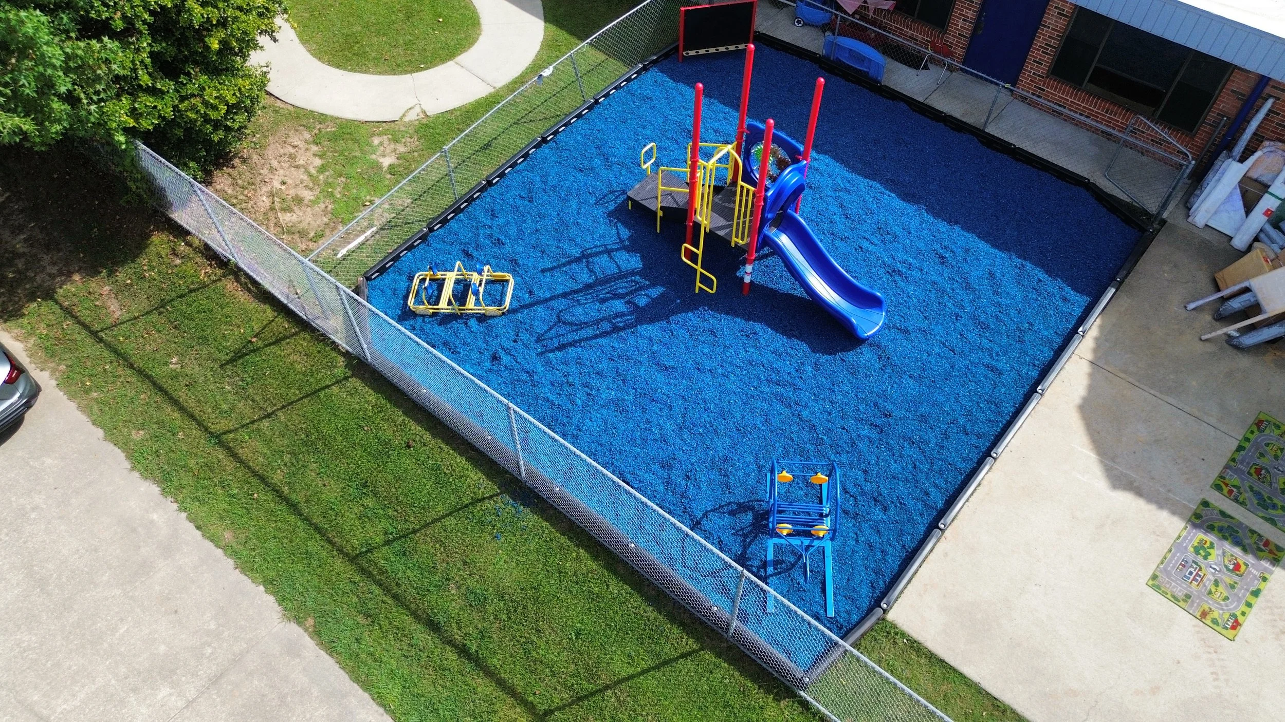 An aerial view of a fenced playground area with a blue rubberized surface, featuring a slide, a climbing structure, and a small platform. Next to the playground is a concrete sidewalk, some grass, a tree, and nearby garage or storage area with some b