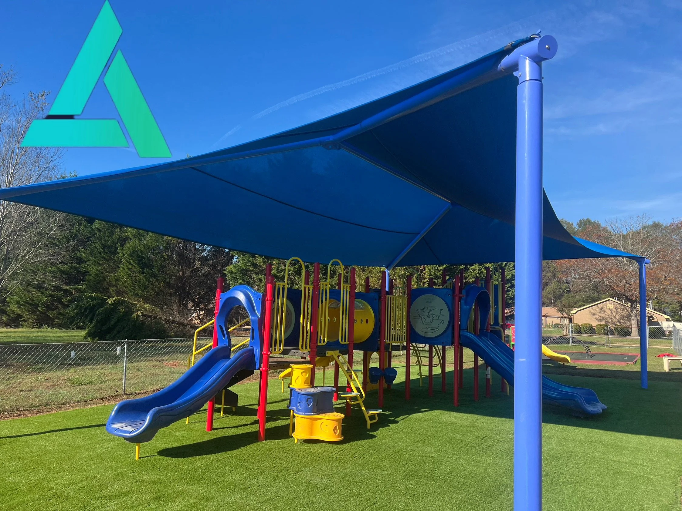 Colorful playground structure with slides, climbing areas, and a shade canopy in a park, surrounded by trees and a chain-link fence.