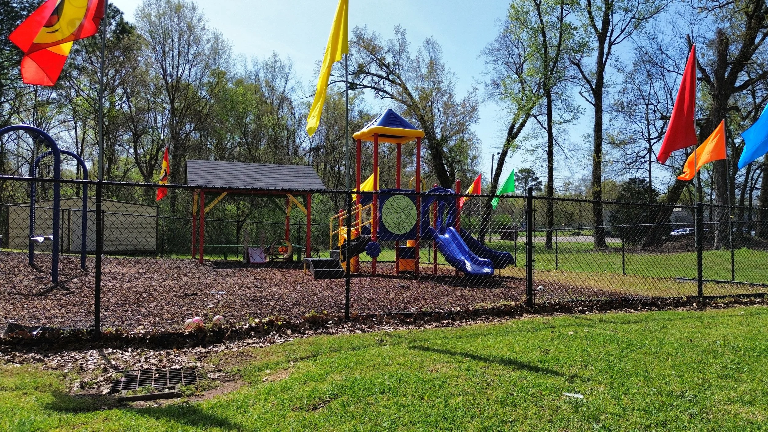Colorful playground with slides and flags, surrounded by trees and a black fence, on a sunny day.