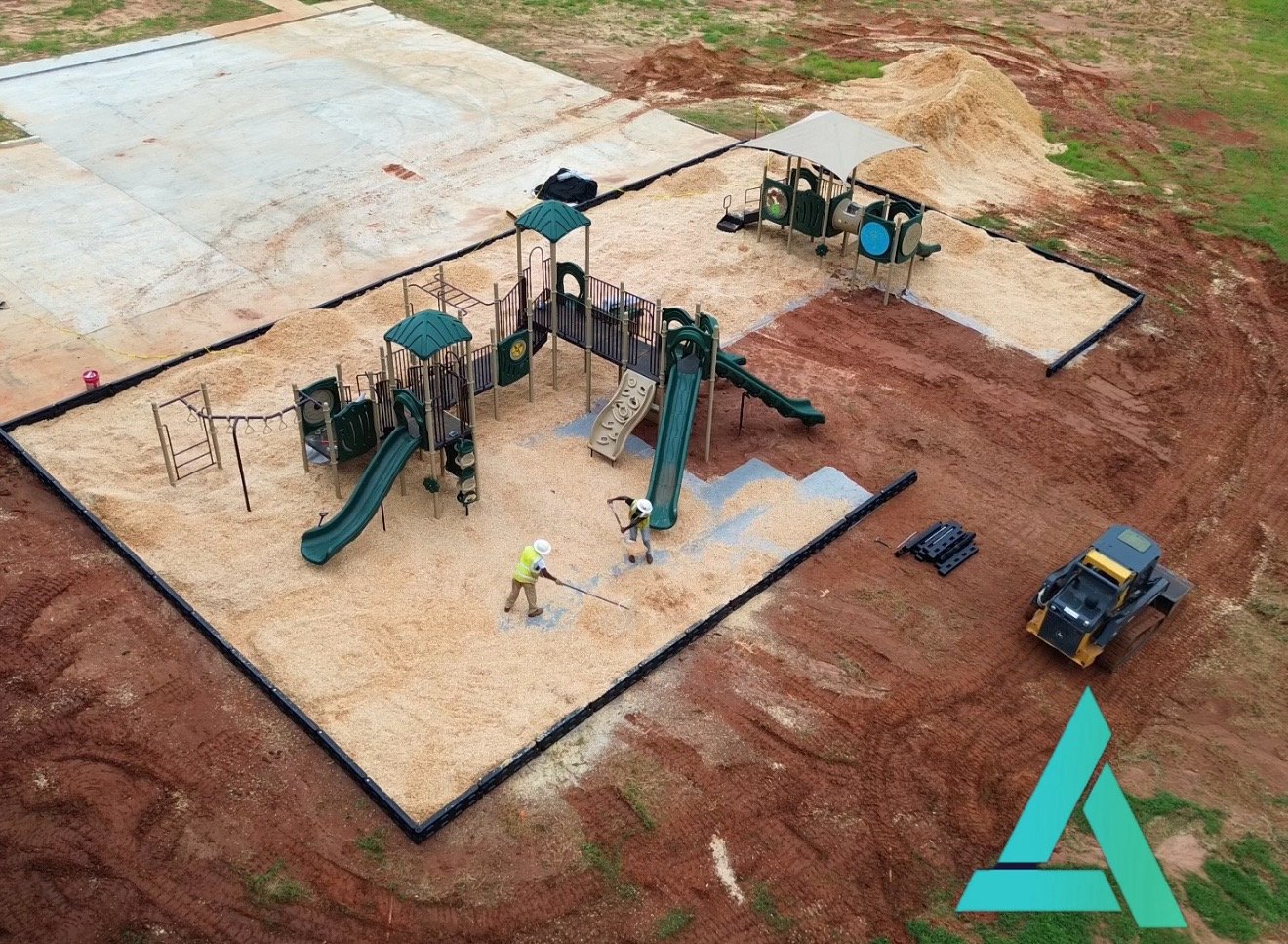 Construction workers installing a playground with slides, climbing structures, and swings inside a sand area, surrounded by red dirt and partially paved ground.