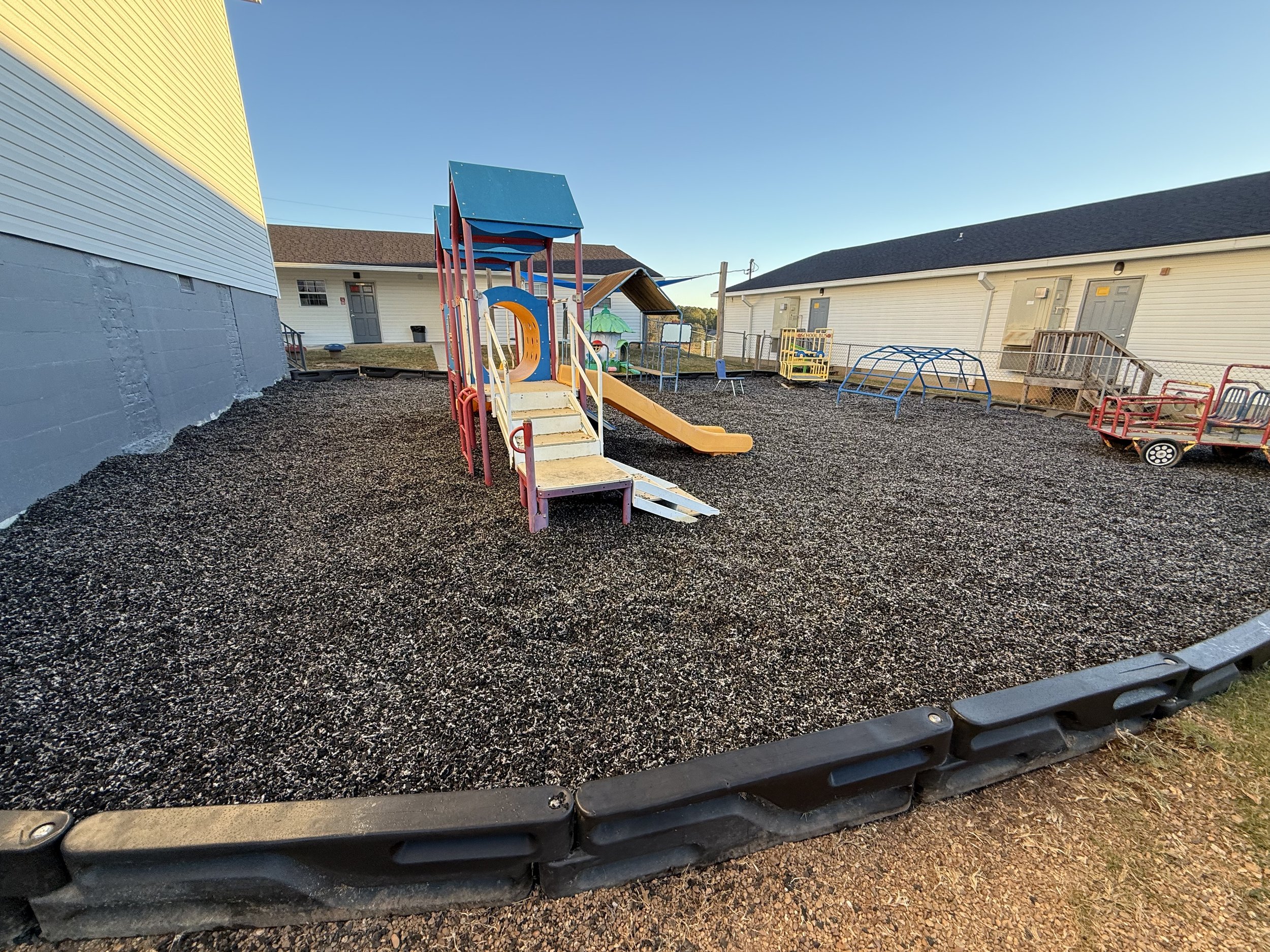 A playground area with various play structures and equipment, surrounded by gravel and located near residential buildings on a clear day.