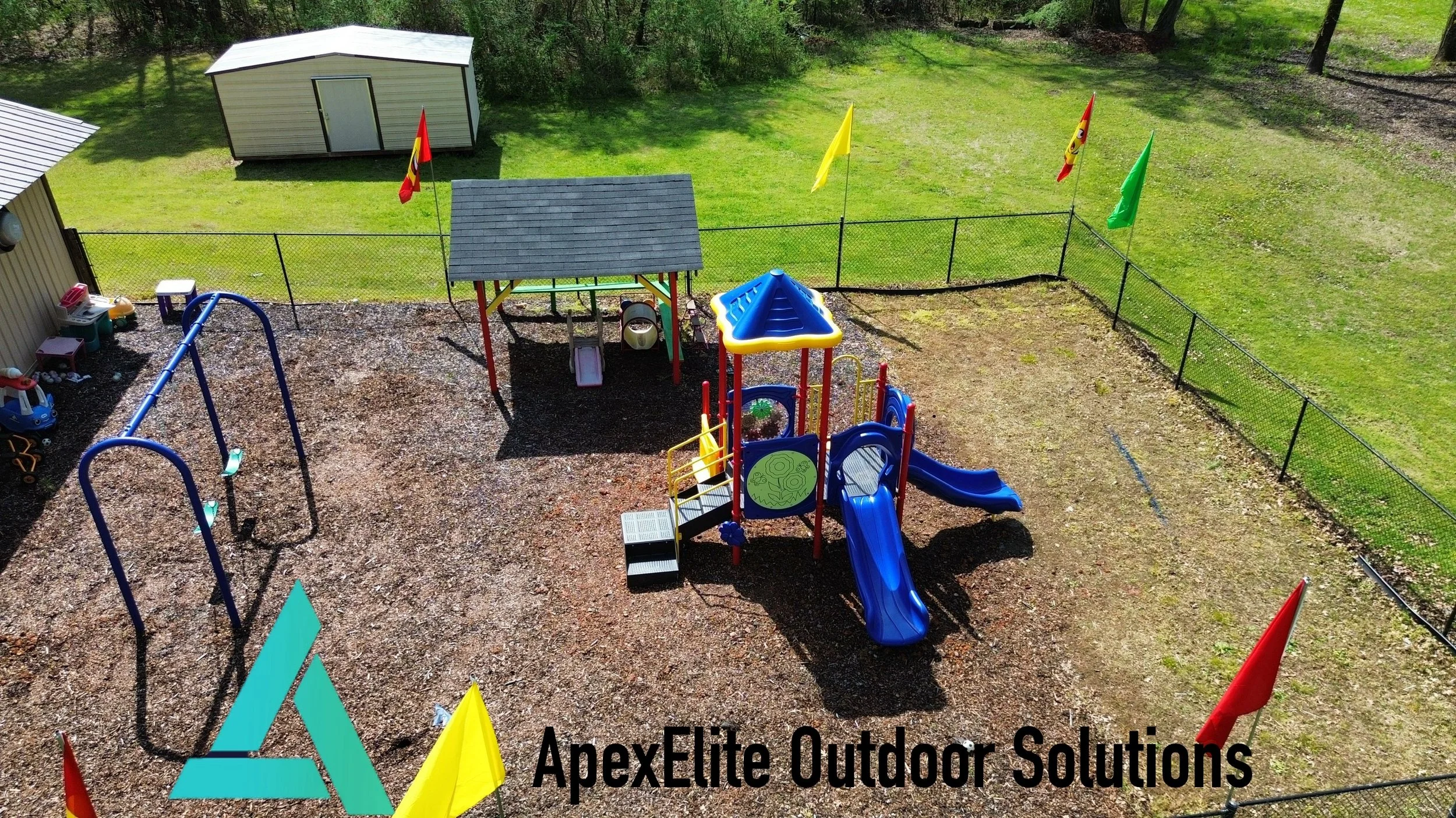 A backyard playground with swings, slides, and a sandbox, surrounded by a black chain-link fence and green grass, with a small shed and trees in the background.
