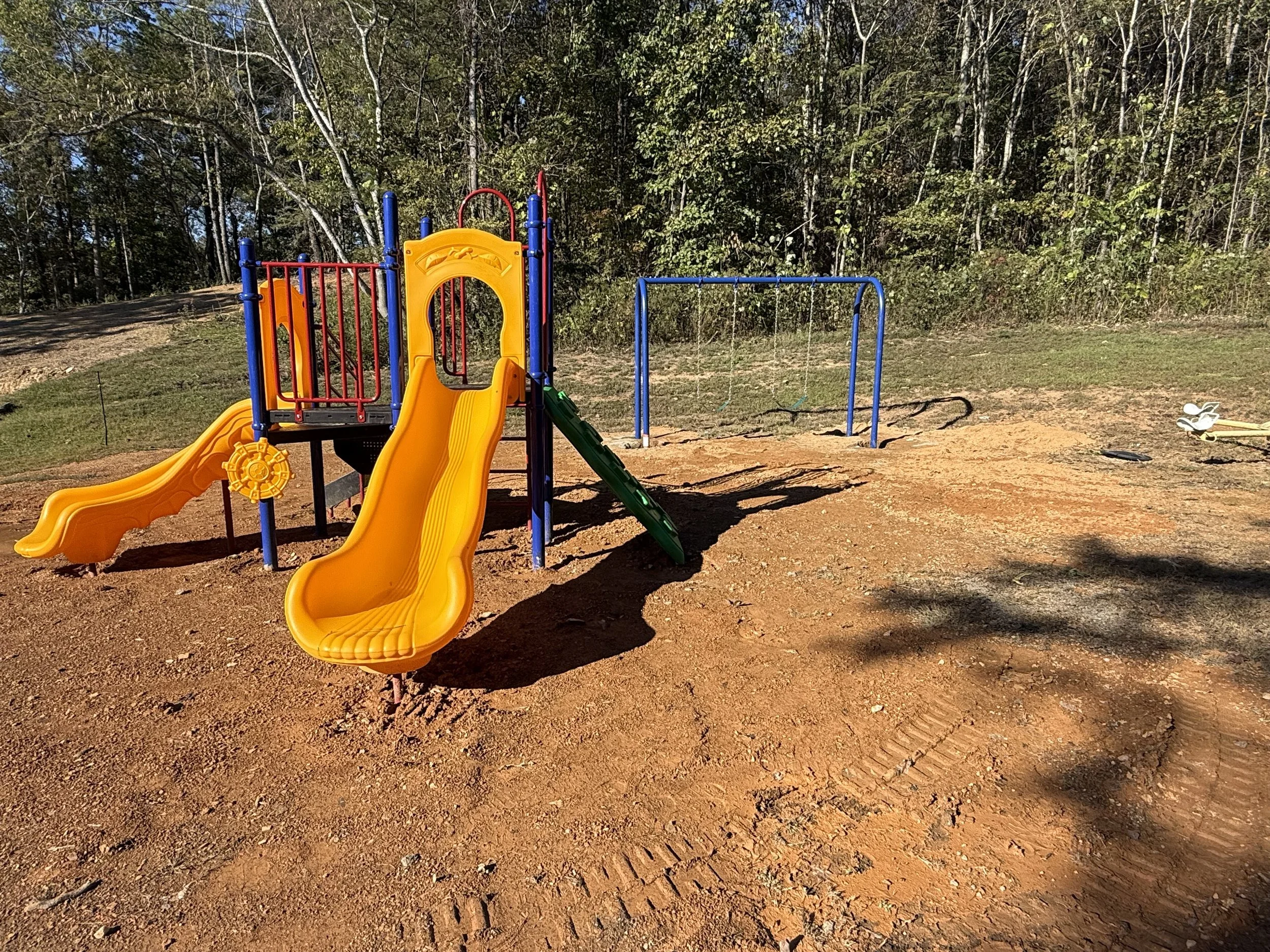 Playground with yellow slide, blue swing set, and green slide, surrounded by dirt and trees in the background.
