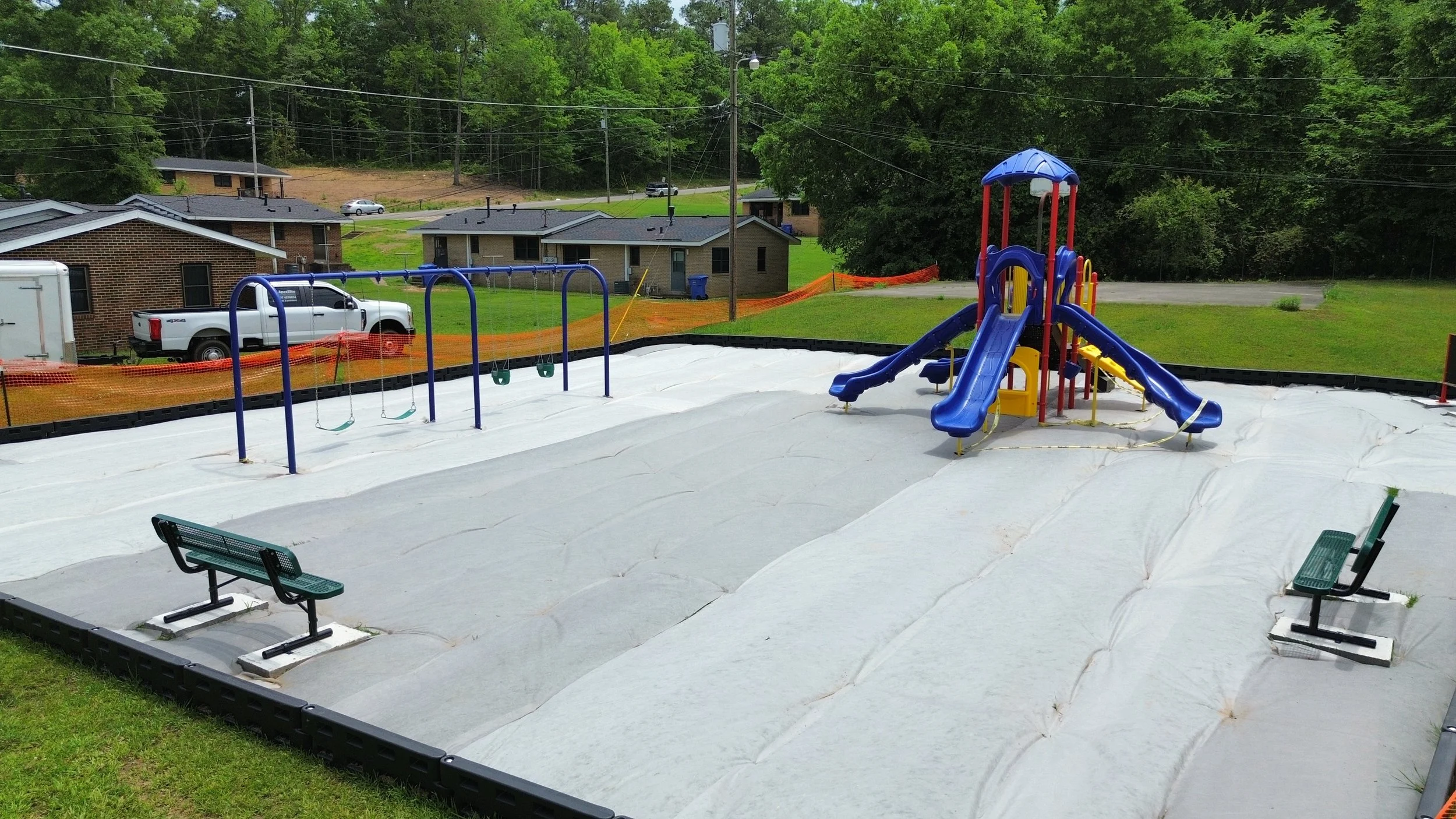 Empty water park with a small blue and red playground slide, swings, and benches, surrounded by green grassy area and trees.