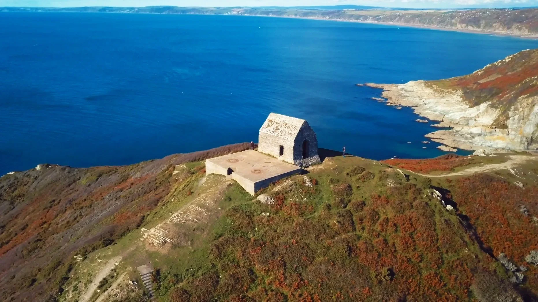 Aerial view of a small stone building on a grassy, rugged hill overlooking a large blue body of water with rocky shores.