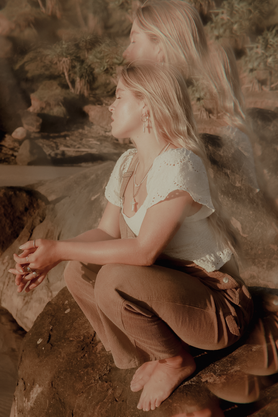 A young woman with blonde hair, wearing a white eyelet blouse and brown pants, is sitting on a rock near water with her eyes closed, and the photo has a double-exposure effect showing her profile and face. She is barefoot and jewelry, including earrings and necklaces, is visible.