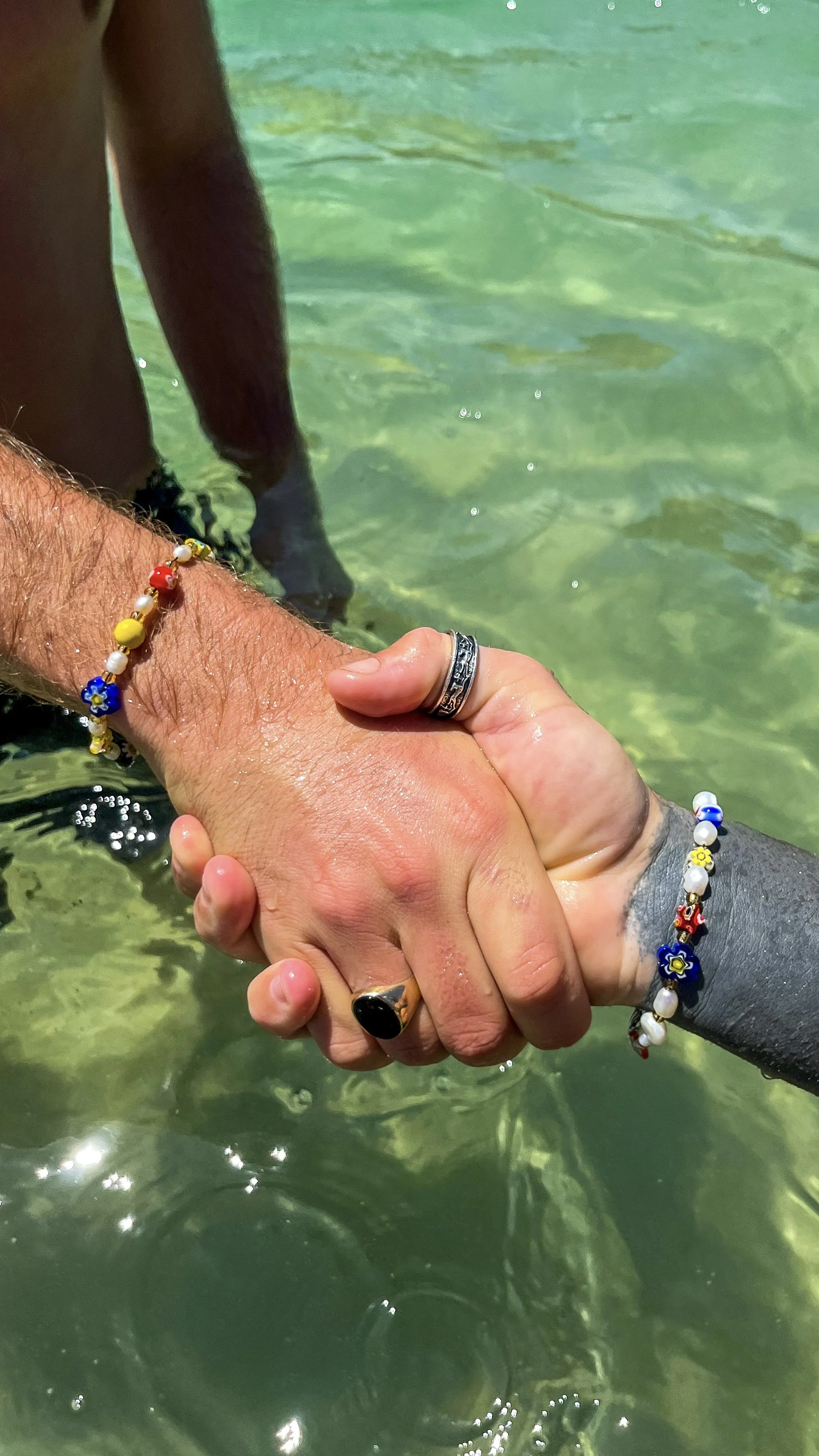 Two people are shaking hands underwater in clear green water, wearing colorful beads and rings.