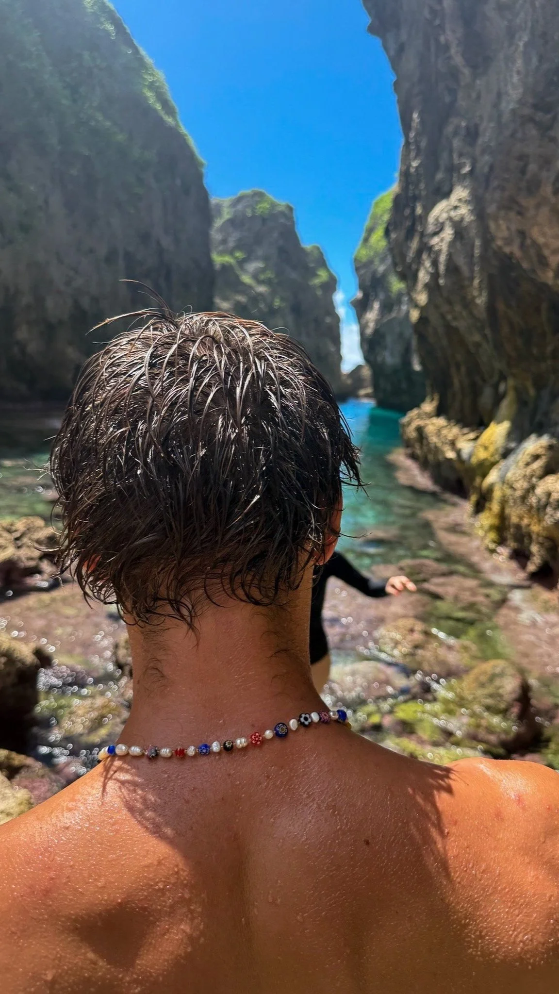 Person with short wet hair and beaded necklace sitting in clear water at a narrow canyon with high rock walls and blue sky.