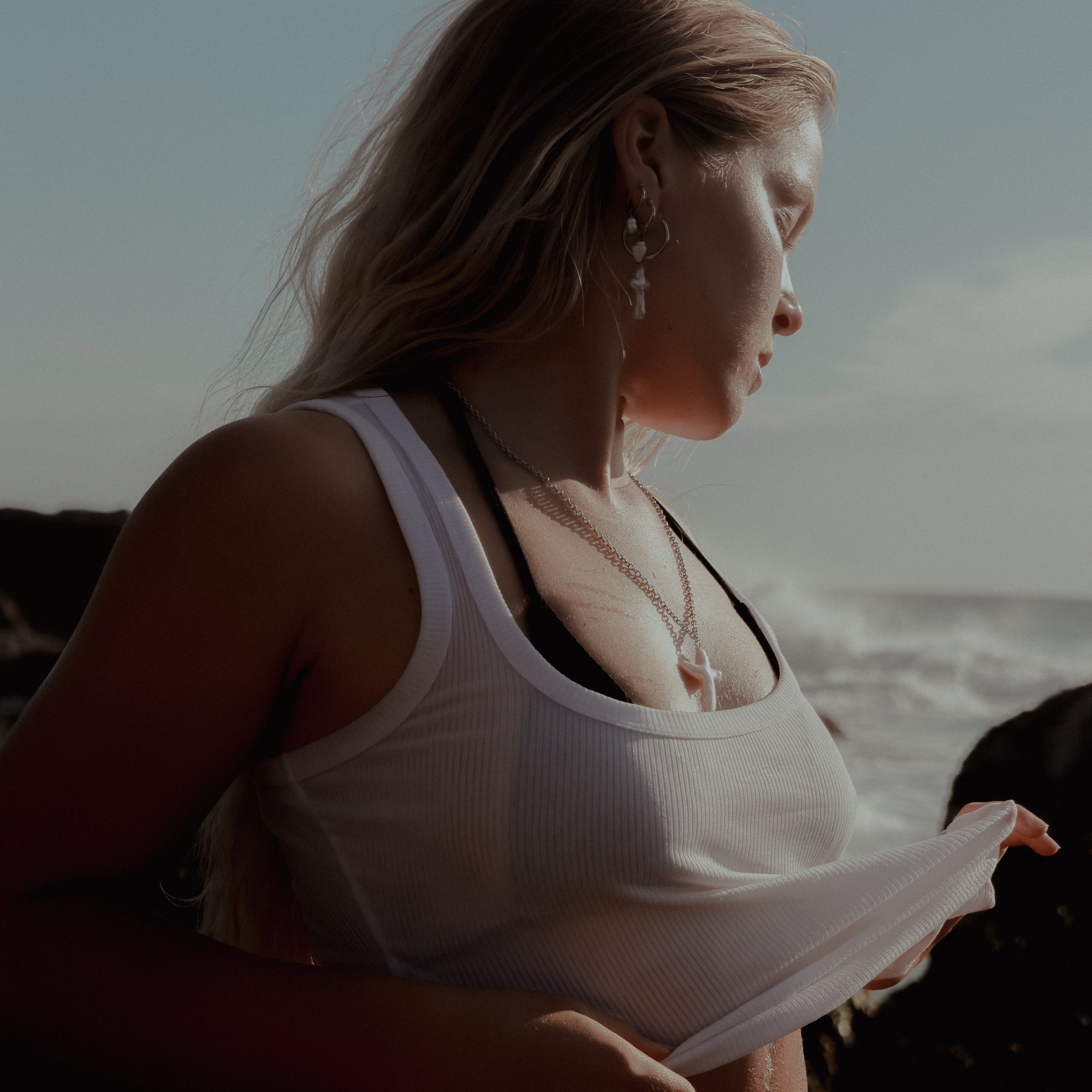 A woman with blonde hair looking down at a white tank top she is lifting, standing near the ocean with rocks and waves in the background.