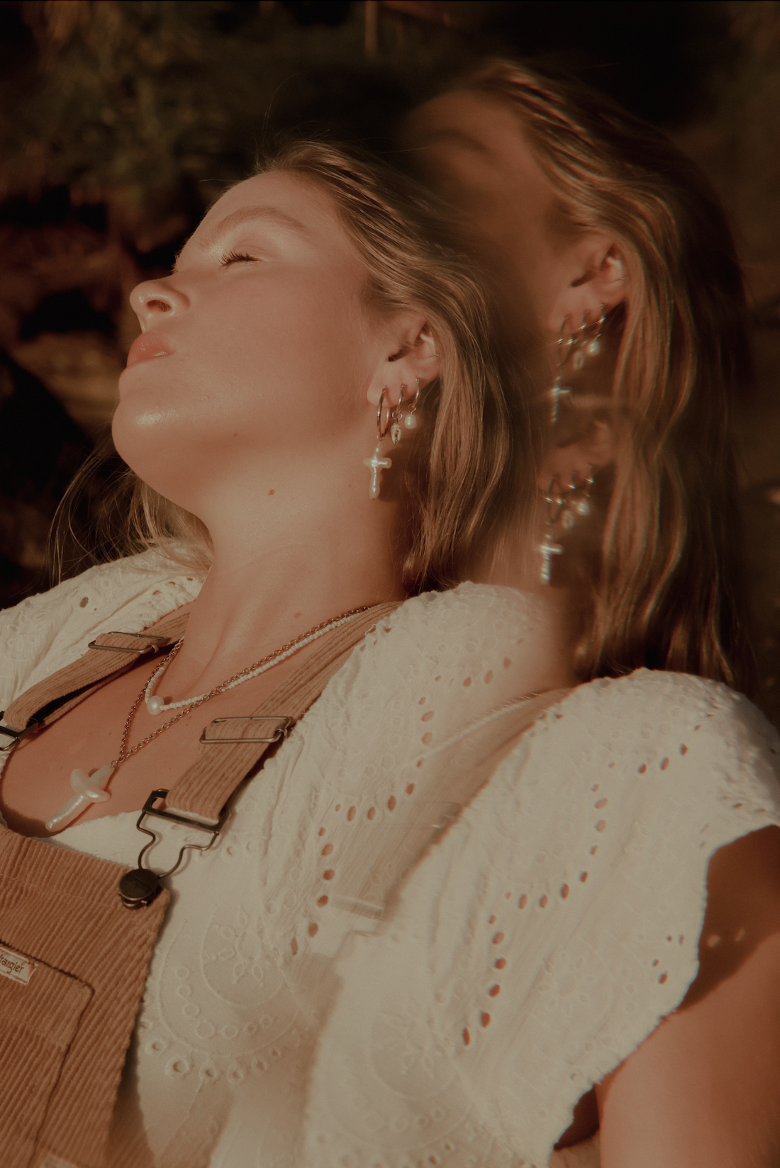 A young woman with light skin, long wavy hair, and multiple earrings, including a cross-shaped earring, smiling and wearing layered necklaces. She has her eyes closed and is lying down outdoors with sunlight highlighting her face, wearing a white embroidered top and tan overalls.
