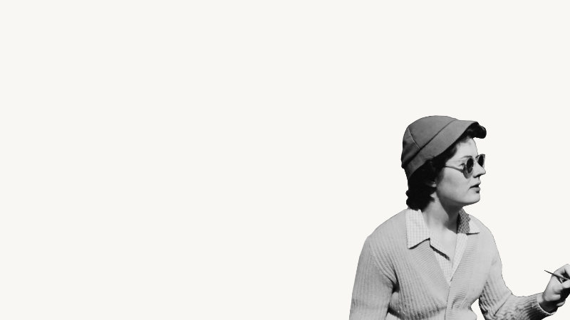 Black and white photo of a woman wearing sunglasses, a hat, and a sweater, sitting sideways and writing or drawing on a piece of paper.