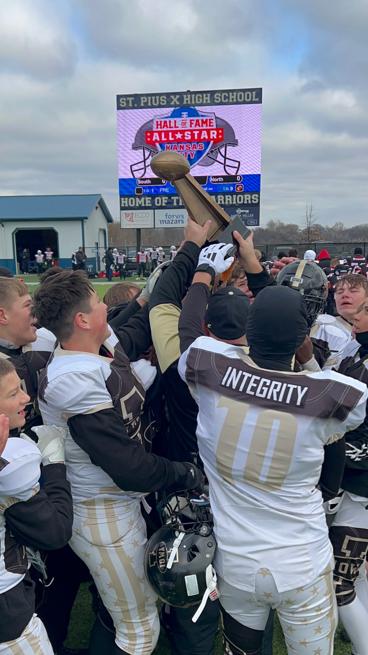 High school football team celebrating victory, holding a trophy, with scoreboard in the background showing they are the home of the Warriors, at St. Pius X High School in Kansas City.