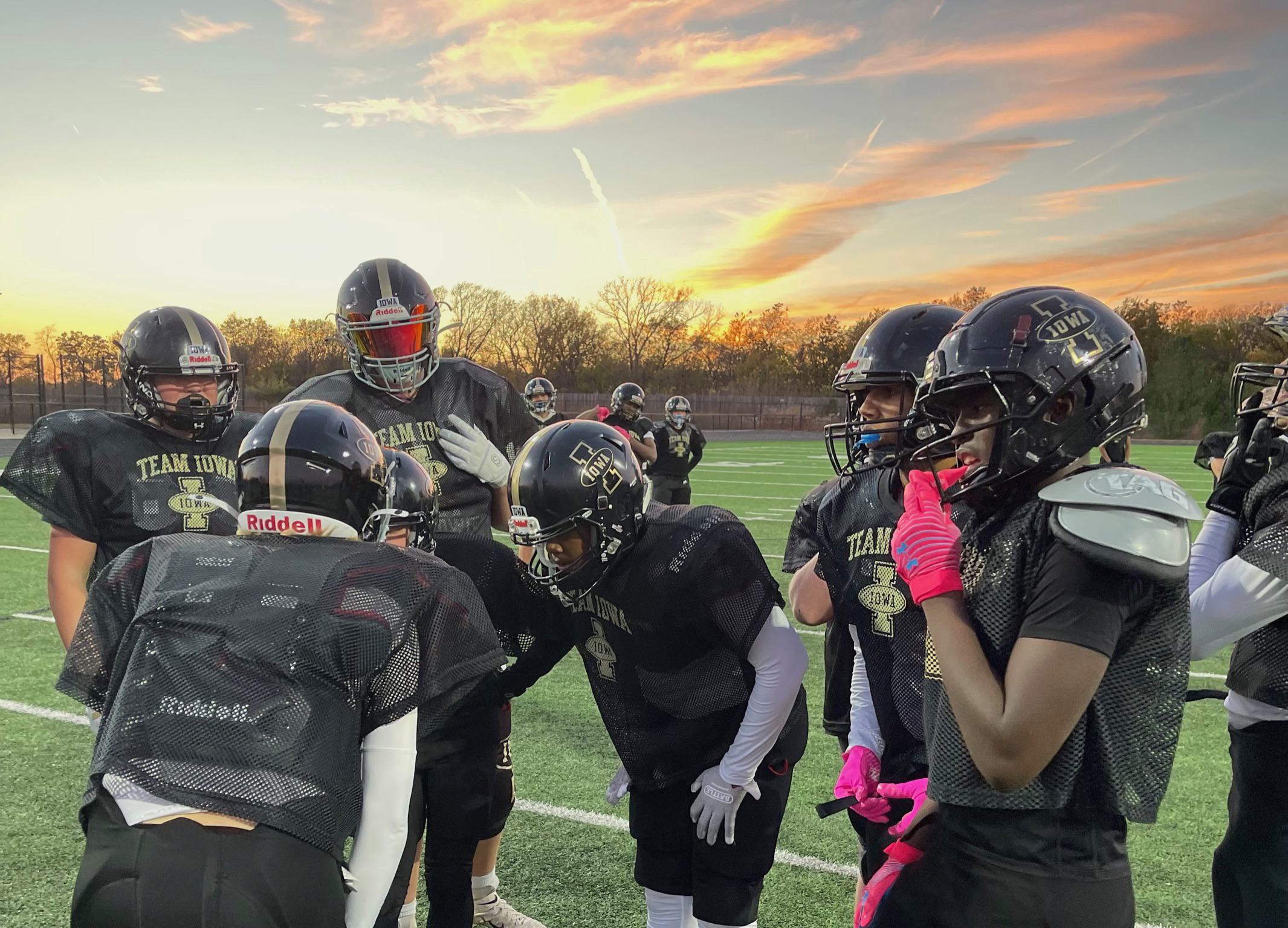 Group of football players in black practice jerseys and helmets gathered on a field during sunset