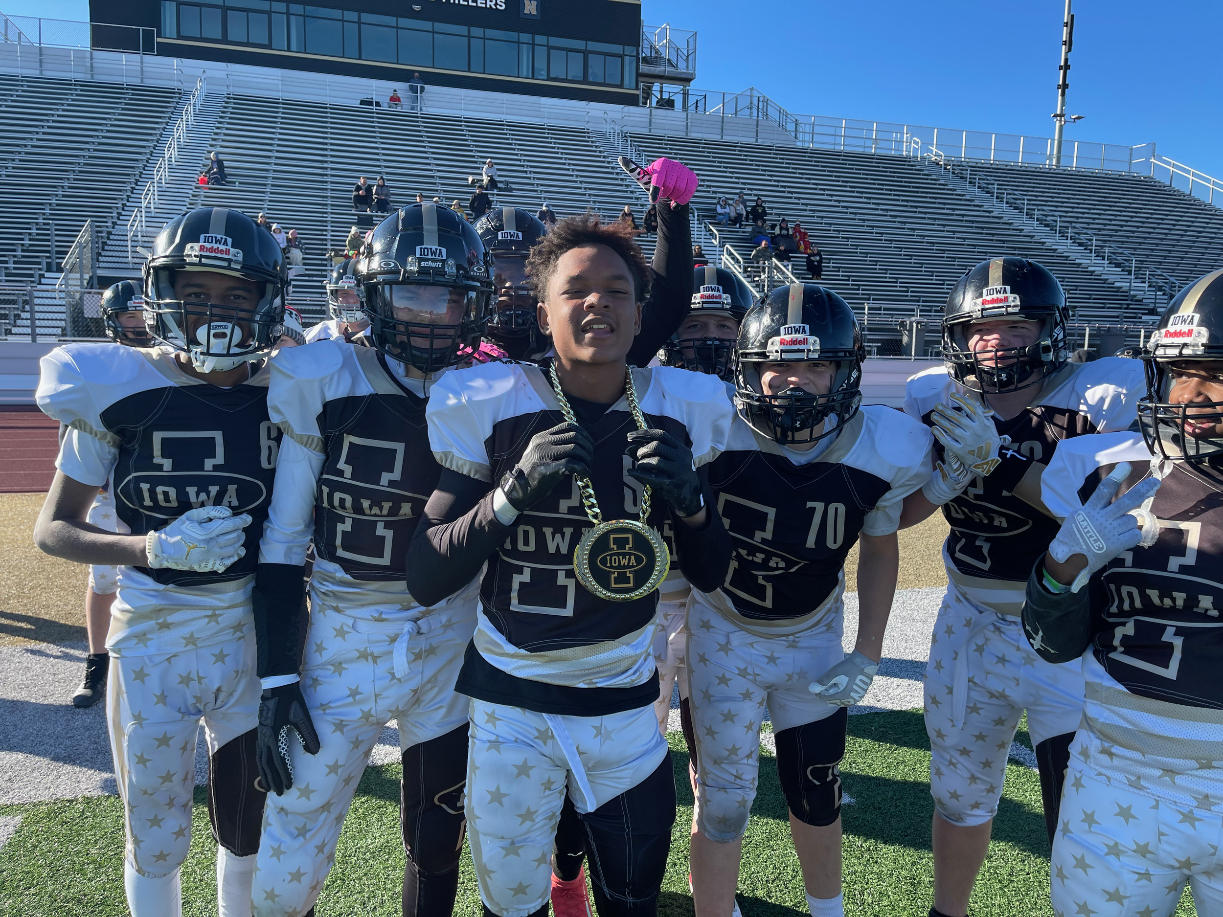 Group of young football players in black and white uniforms with Iowa logos, some wearing helmets, celebrating on the field with a coach or team member holding a medal around his neck, with empty stadium bleachers and a few spectators in the background.
