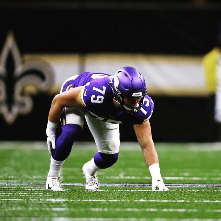 A football player in purple and white uniform crouching on the field, preparing for the play.