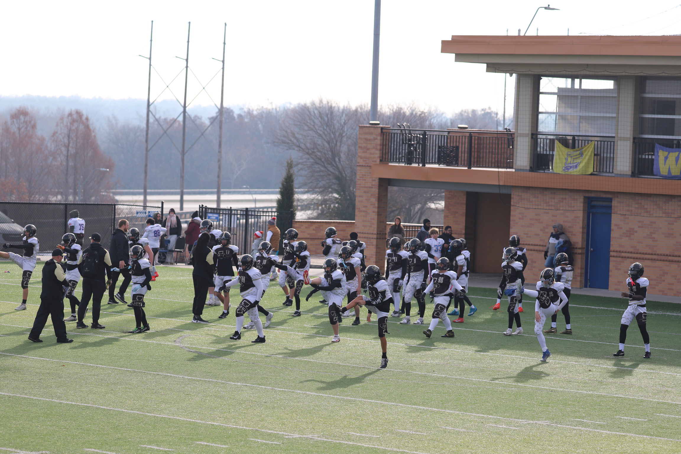 Youth football team practicing on the field with coaches, all wearing helmets and uniforms, in front of a brick building with a balcony and banners.