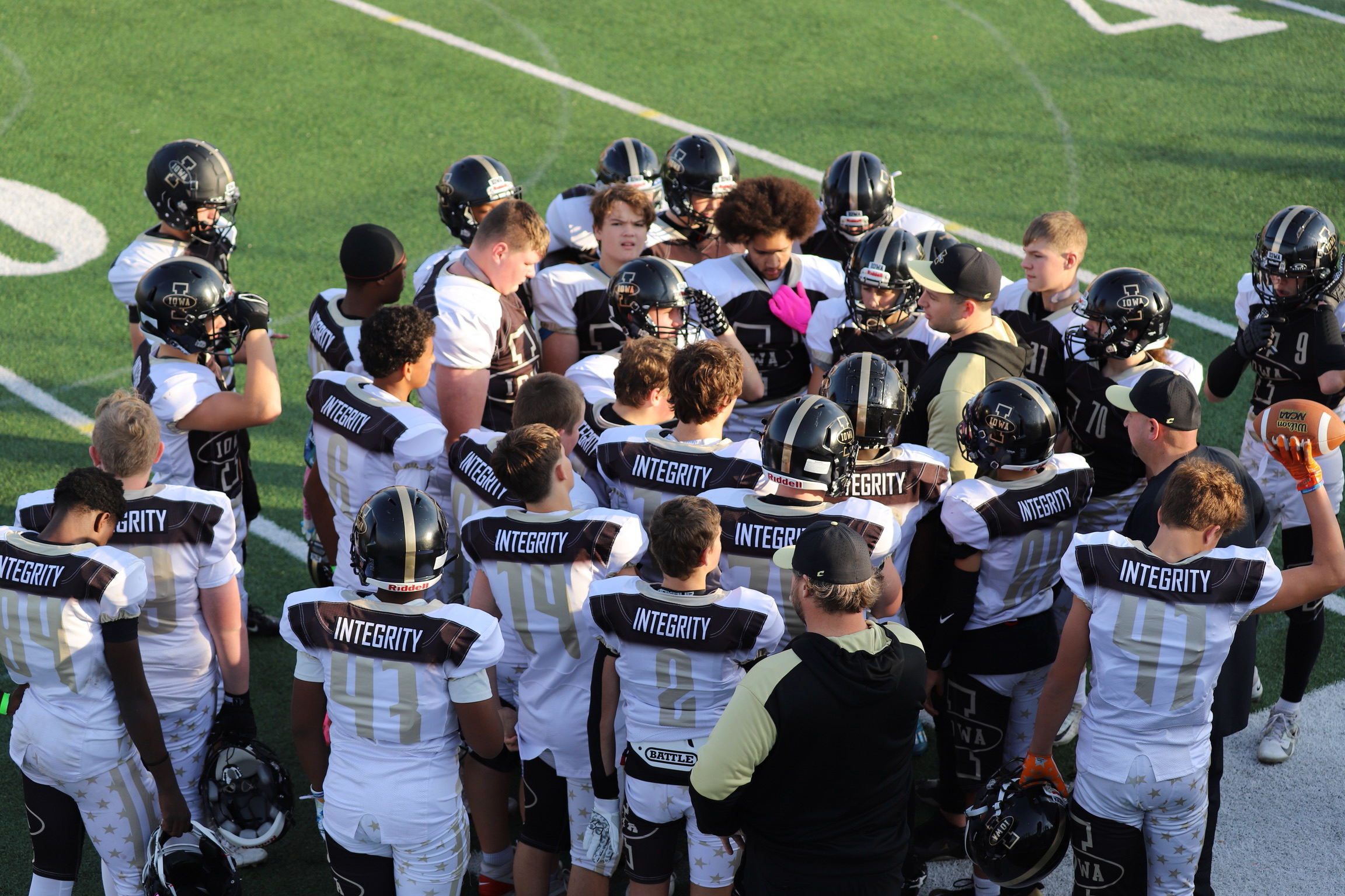 A youth football team in black and white uniforms with the word 'INTEGRITY' on the jerseys, gathered on a football field for a huddle during a game or practice.