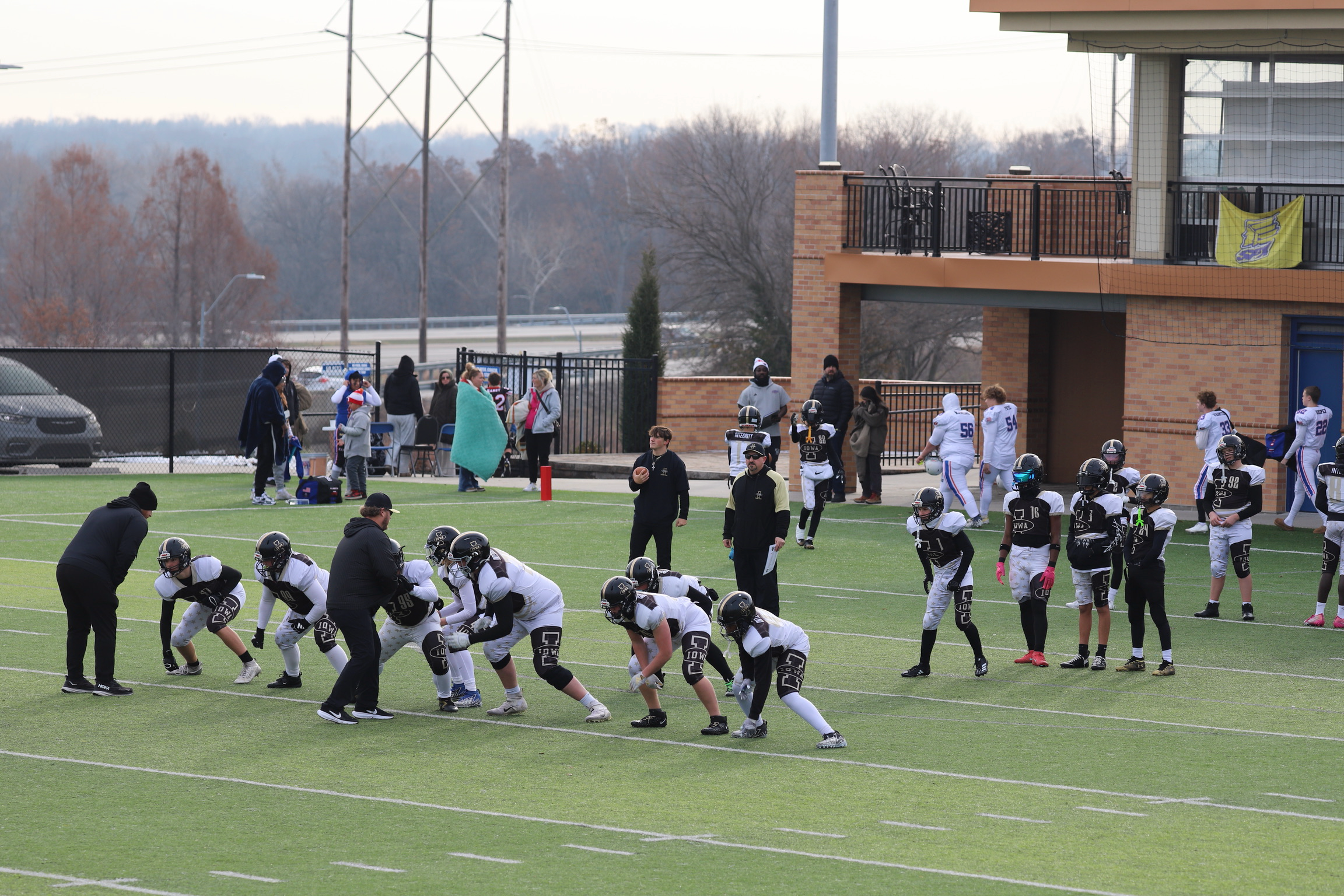 Youth football team practicing on the field, with coaches instructing, and spectators in the background, during a cloudy day.