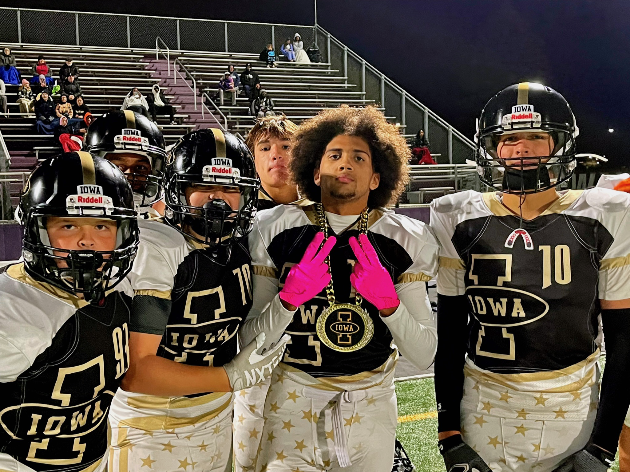 Football team in black and white jerseys with gold stars celebrating on field at night, with spectators in stands behind.
