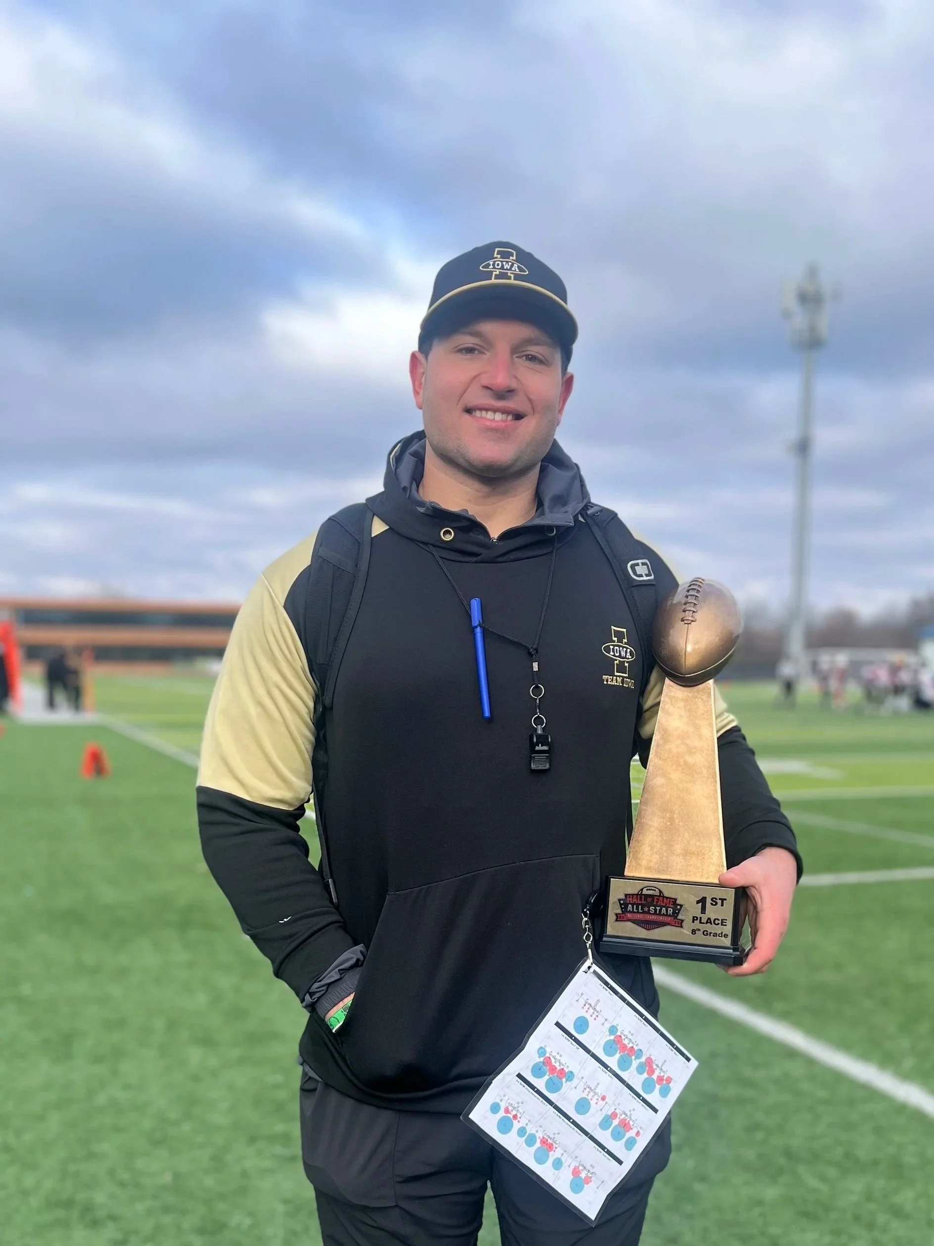 A young man stands on a football field, holding a trophy for first place in an 8th grade all-star game. He is wearing a black and gold sports jacket, a black cap with Iowa and football logos, and a backpack. The sky is cloudy, and there are people and equipment in the background.