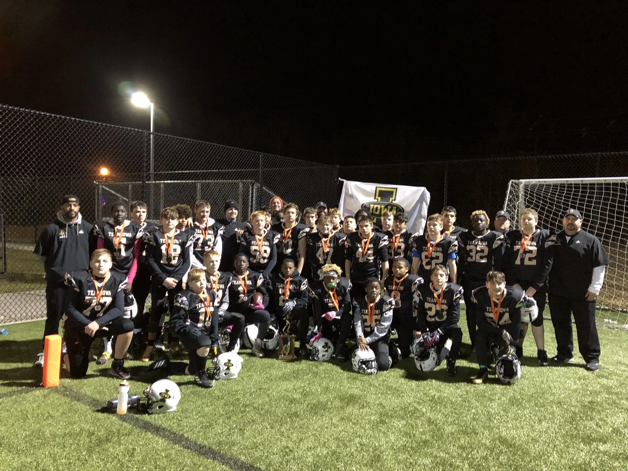 Youth football team in black uniforms with orange medals, posing for a team photo on a well-lit football field at night, with coaches and a banner.