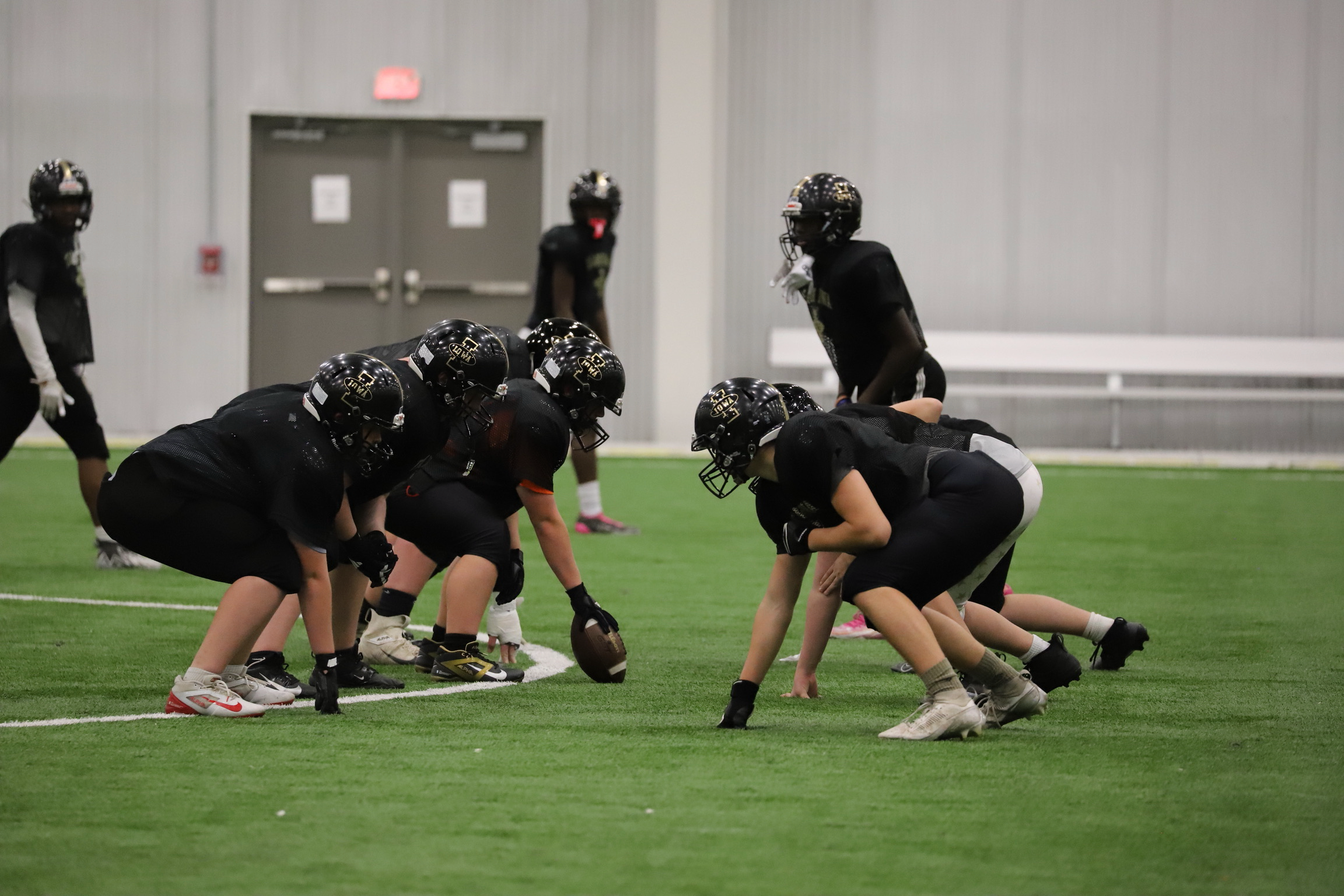 Youth football team practicing on indoor field, players in black uniforms and helmets lined up in formation, preparing for play.