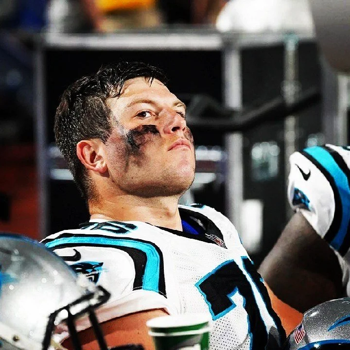 Close-up of a football player with black eye makeup on his face, wearing a white football jersey with blue and black accents, sitting on a bench in a locker room.