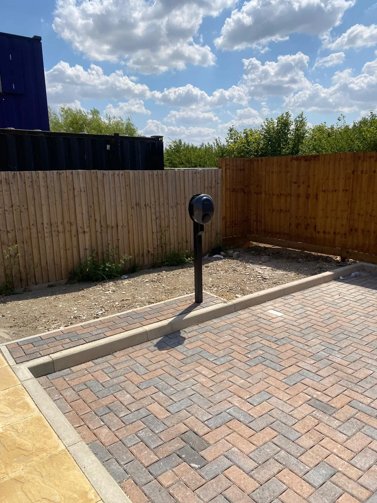 Public parking space with a coin-operated parking meter, brick pavers, a wooden privacy fence, and blue sky with white clouds.