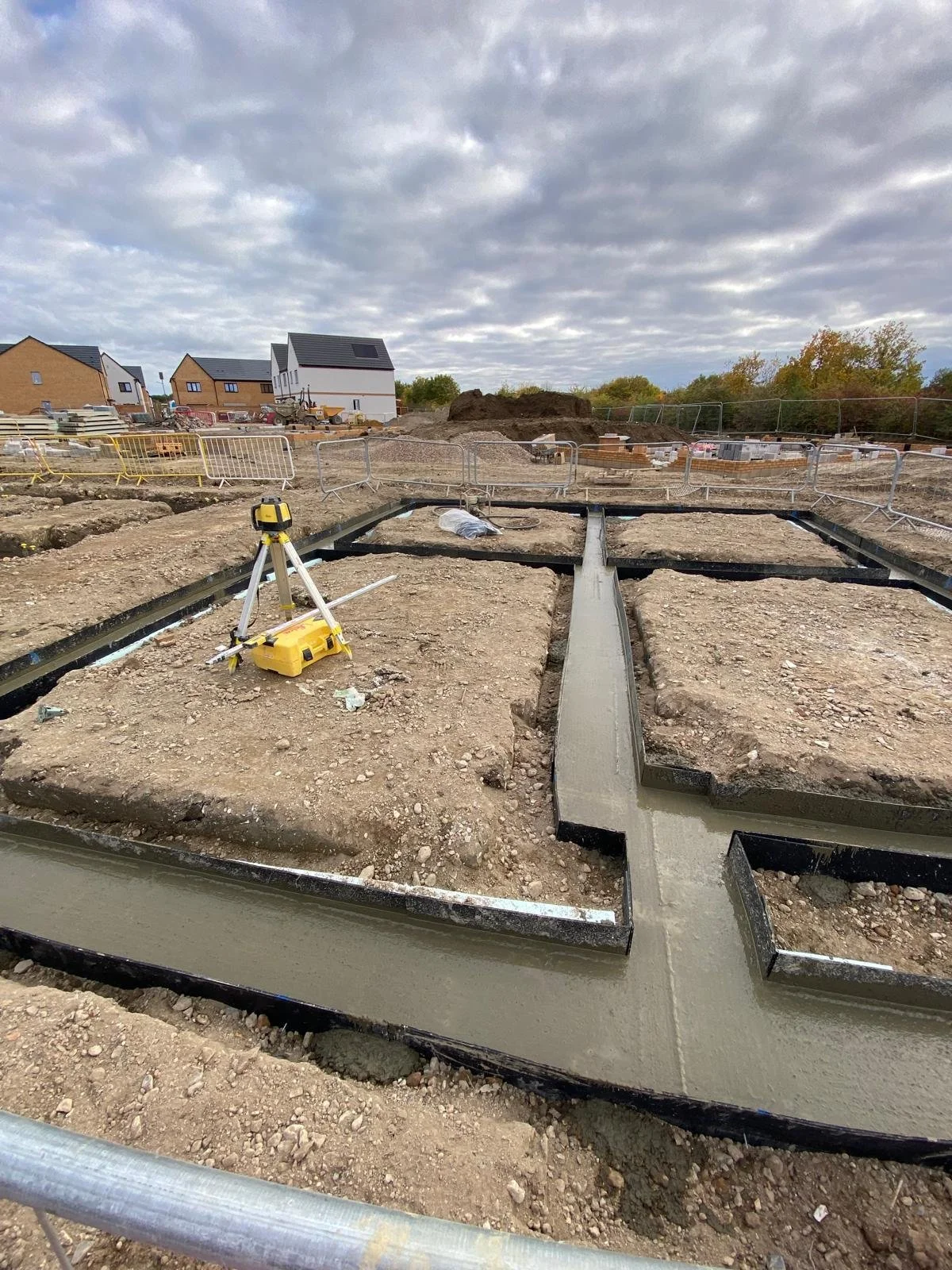 Construction site with freshly poured concrete in trenches, survey equipment, and partially built houses under a cloudy sky.