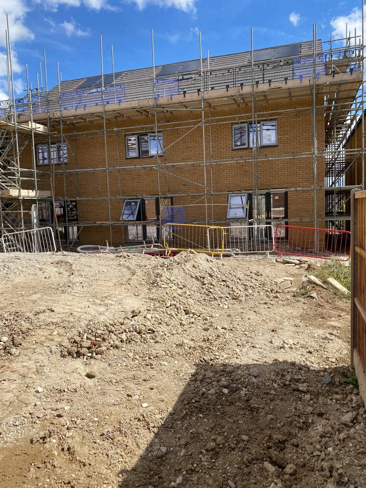 Construction site with a multi-story brick building surrounded by metal scaffolding and safety barriers, with piles of dirt in the foreground and a blue sky with clouds overhead.