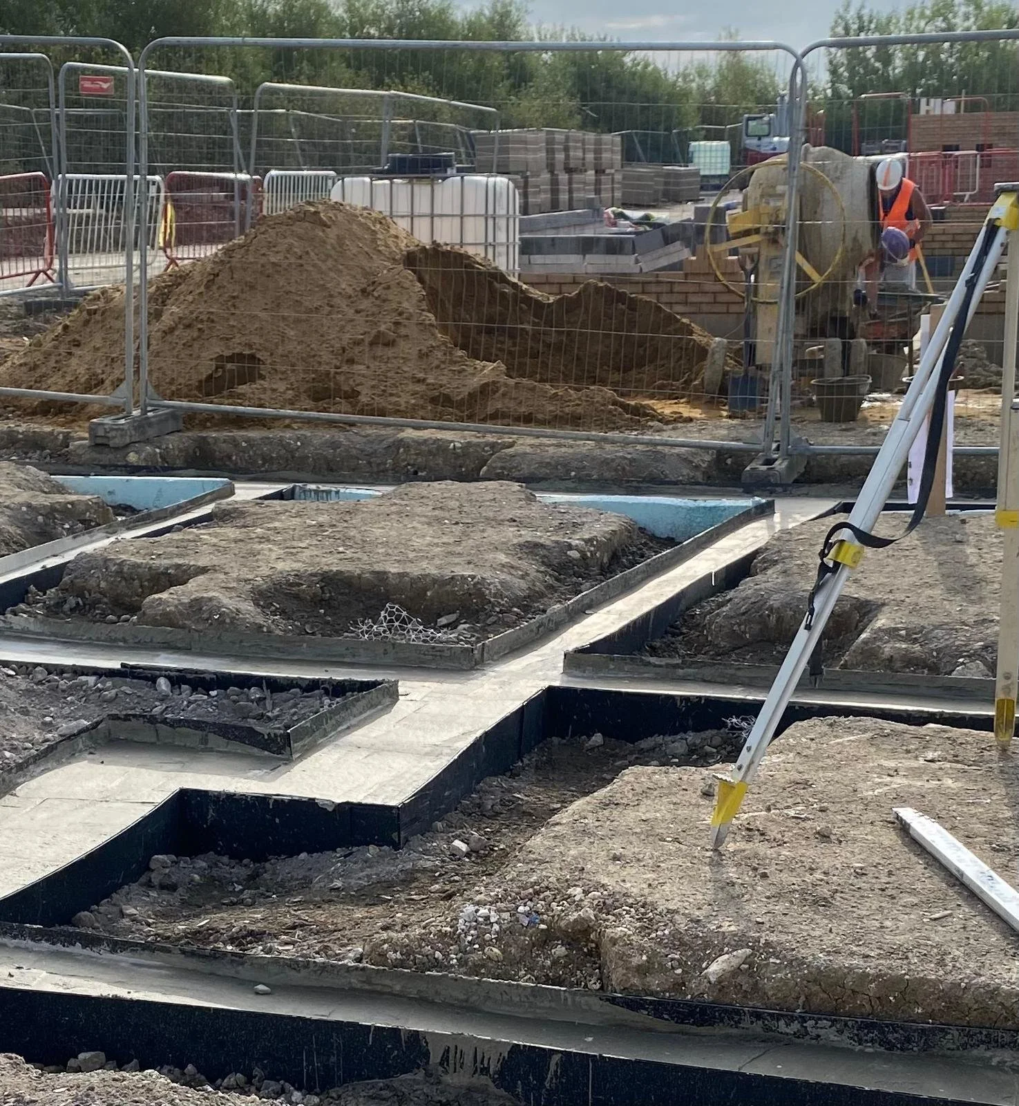 Construction site with workers, digging and laying foundations for a building, surrounded by construction fences, with piles of dirt, construction materials, and equipment.