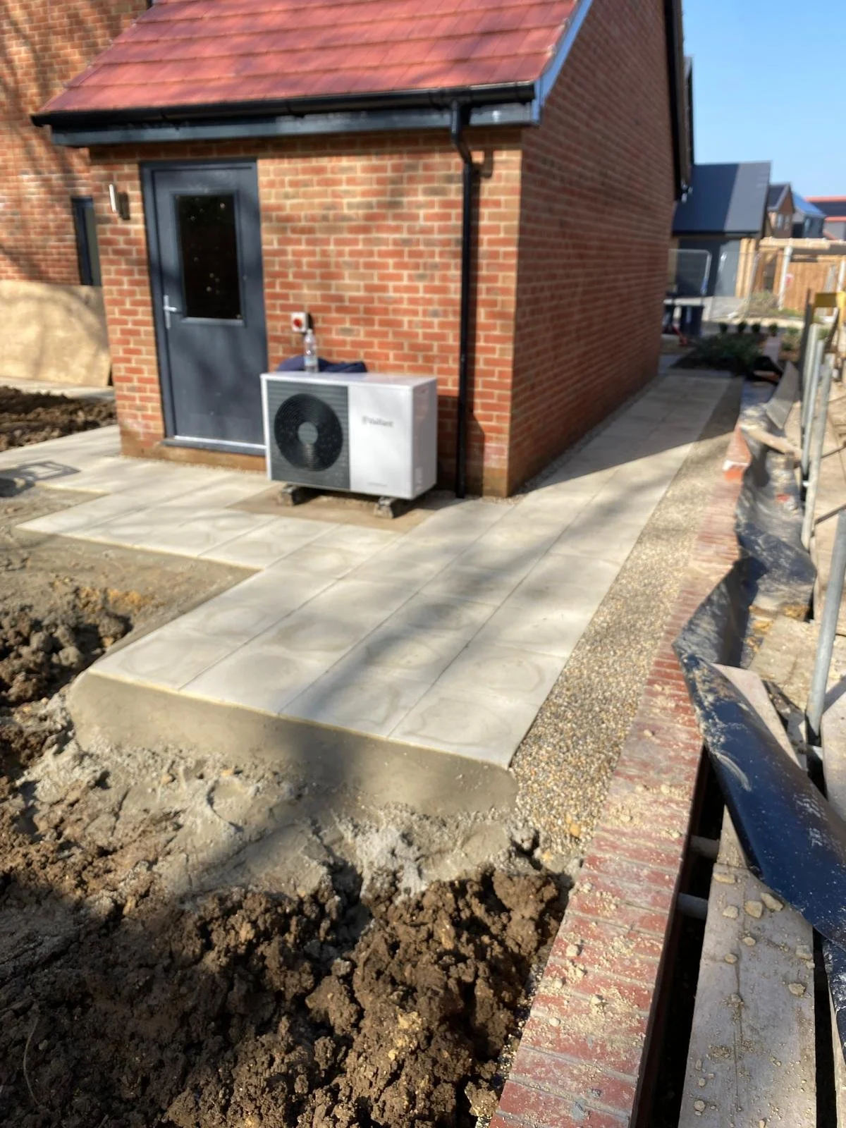 Newly paved walkway and small patio area outside a brick house with a black door, air conditioning unit, and construction materials.