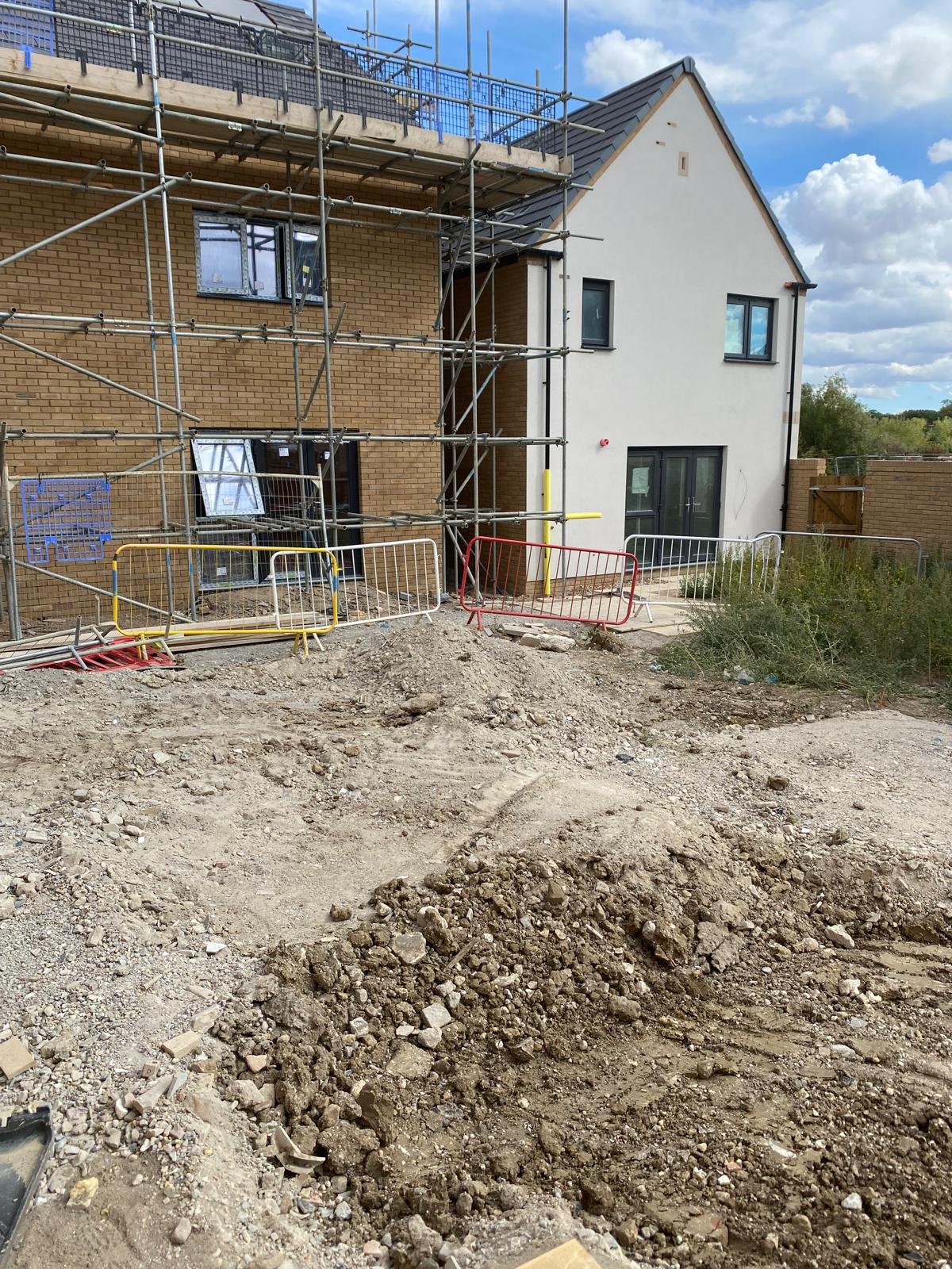 Construction site in front of a residential building with scaffolding and barriers, showing disturbed dirt and gravel ground.