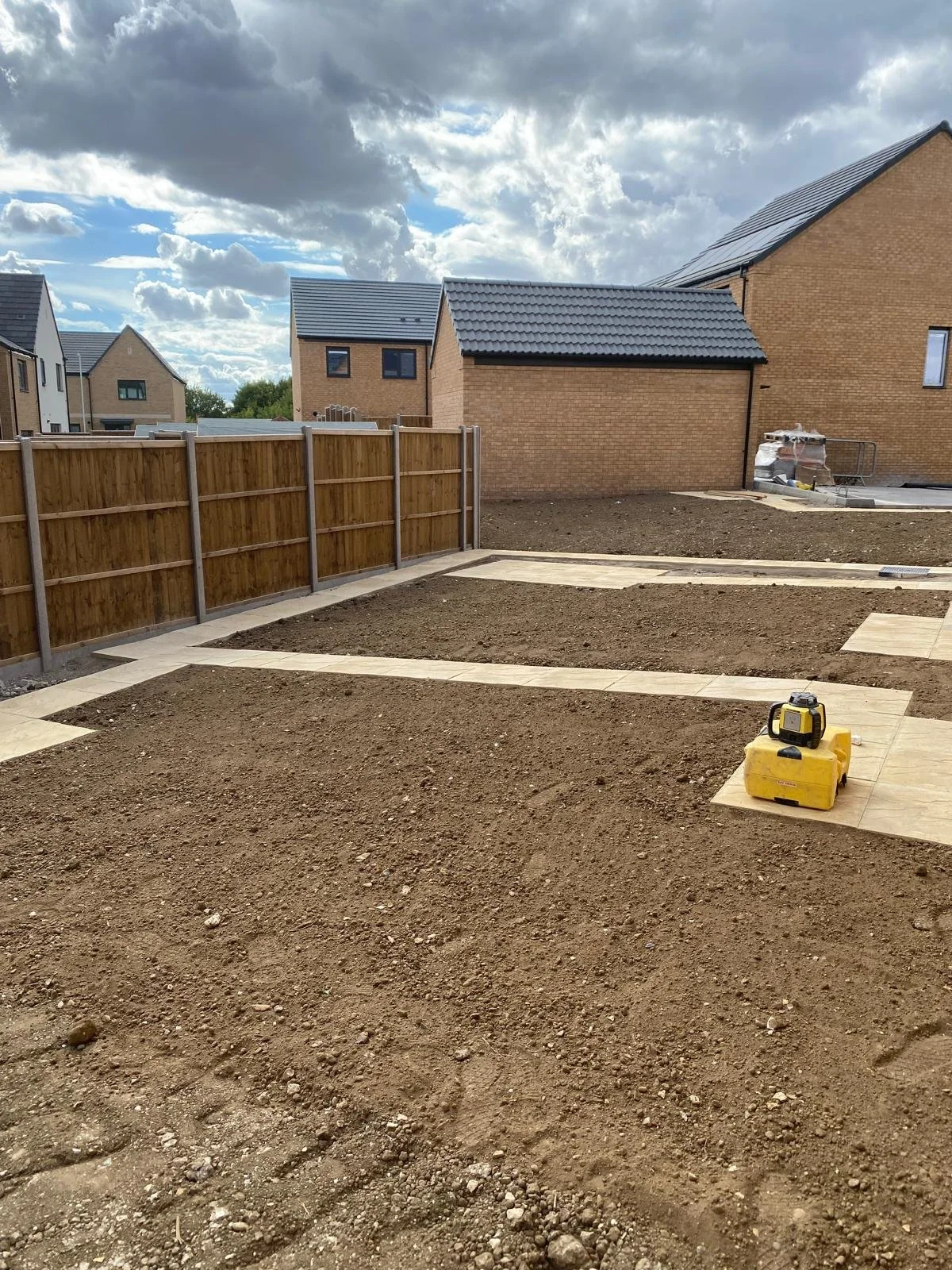 A backyard under construction with dirt ground, recently laid paving stones forming pathways, a yellow laser level tool on one of the stones, a wooden fence on the left, and neighboring houses with brick walls and dark roofs in the background, under 