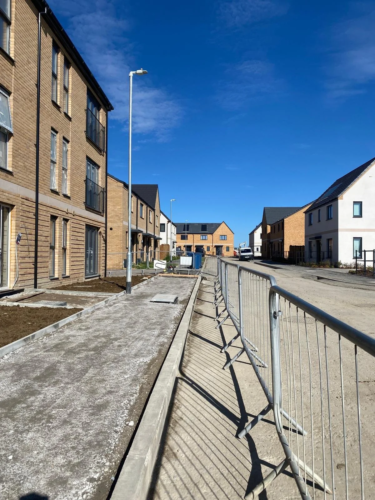 Construction site on a residential street with partially paved sidewalks, metal barriers, brick and white houses, a clear blue sky, and street lamps.