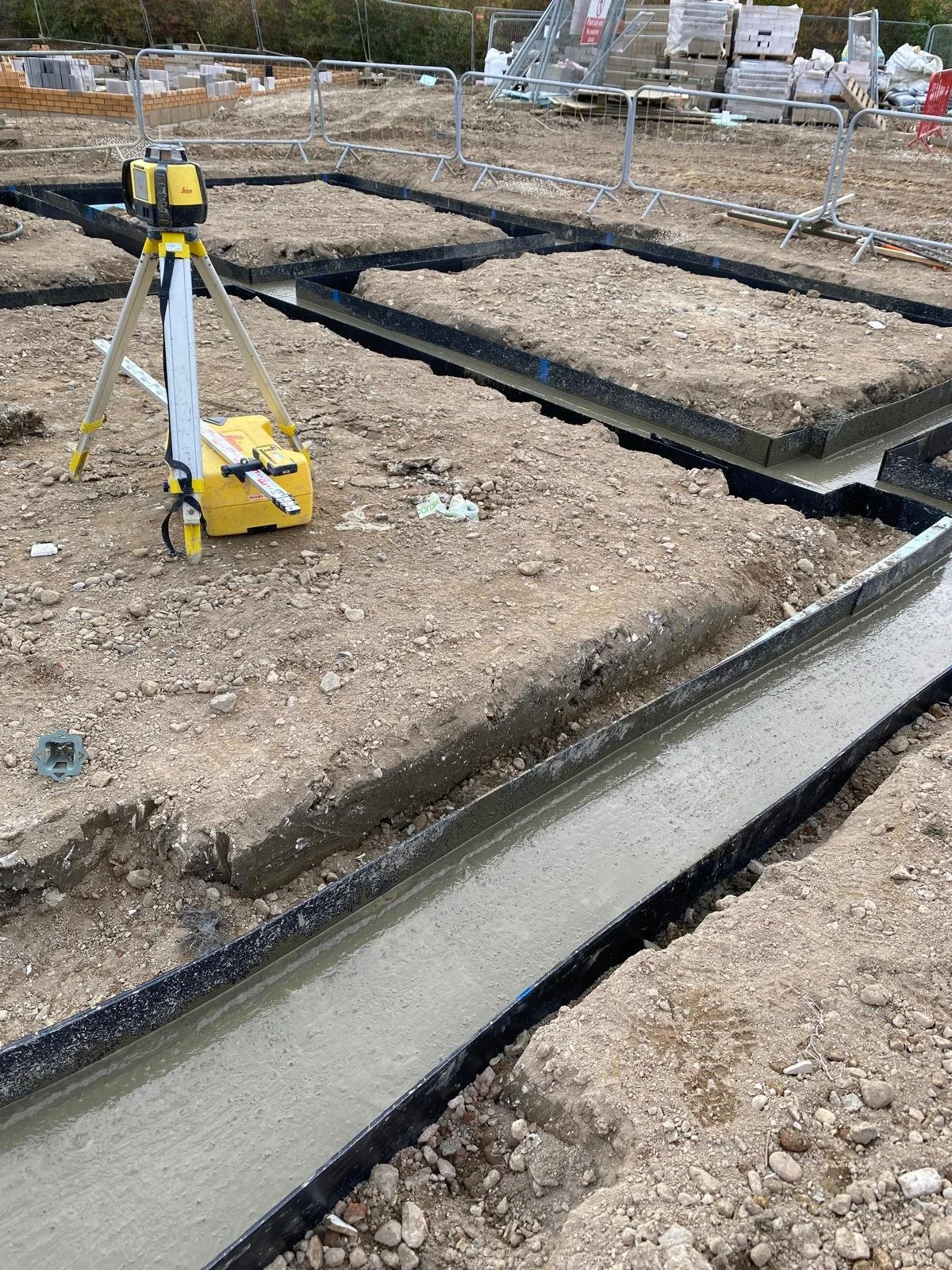 Construction site with concrete foundation and a level on a tripod, surrounded by dirt, metal barriers, and building materials.