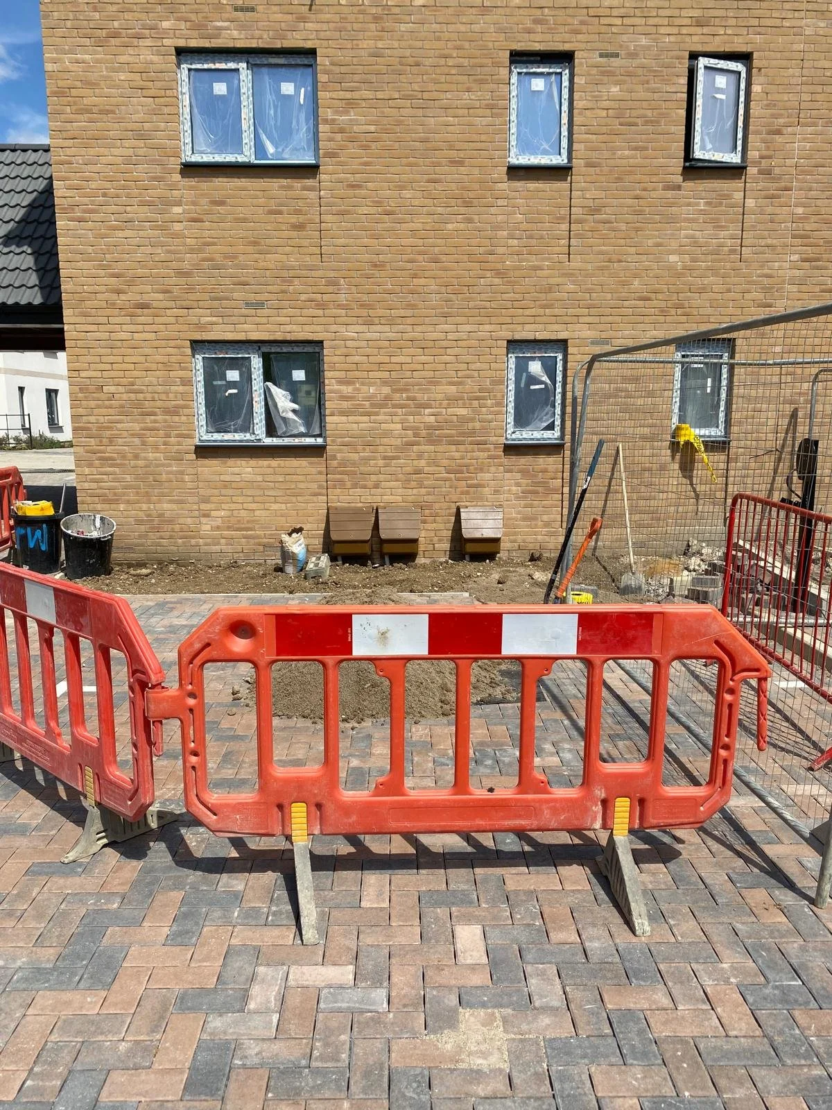 Construction site with orange barricades set up on a brick-paved sidewalk, in front of a building with a brick exterior and six windows, some of which are covered or have protective film.