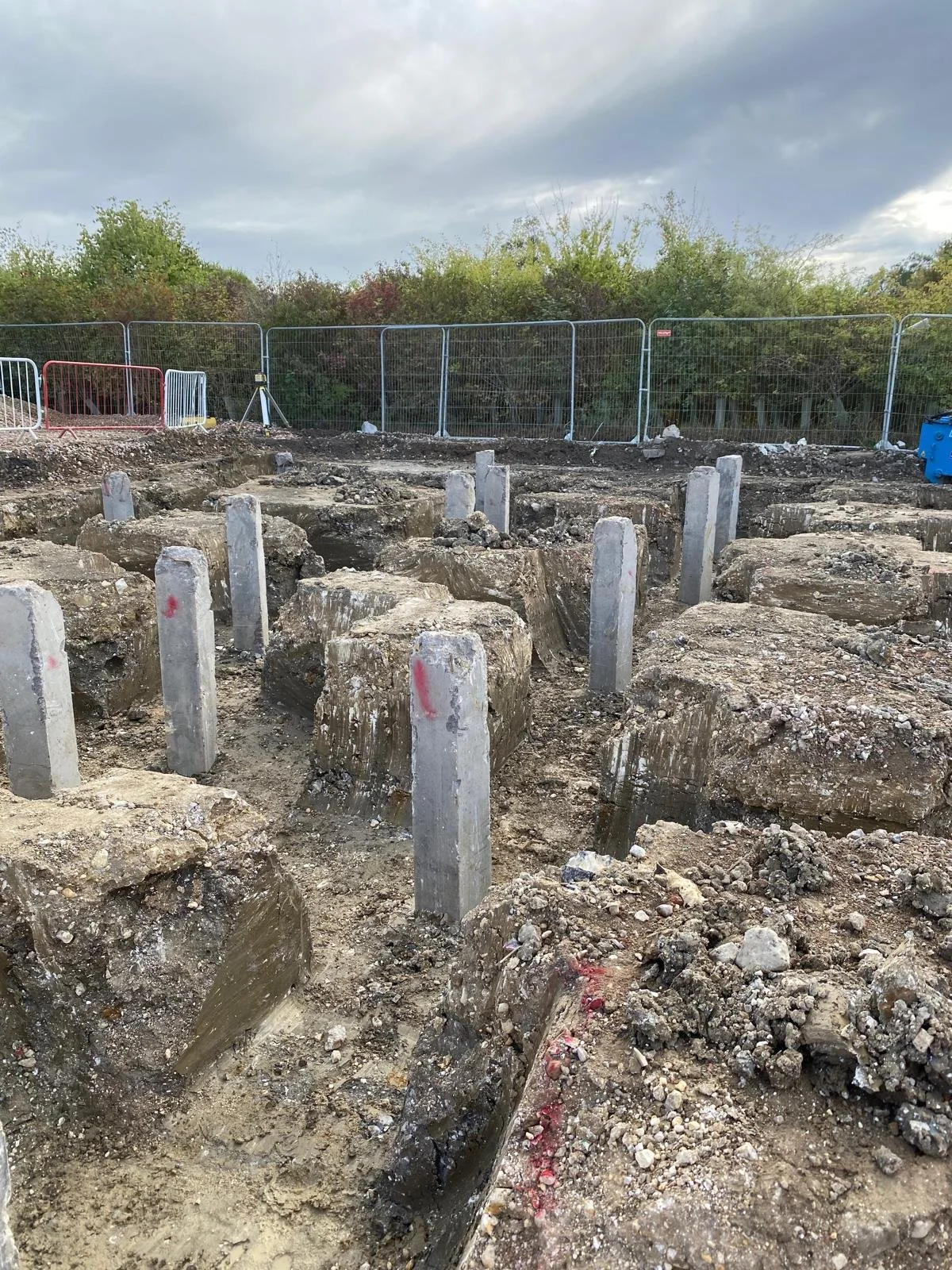 Construction site with exposed foundation and concrete pillars, surrounded by metal fencing and trees in the background.