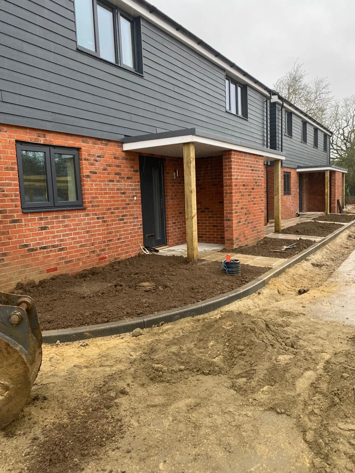 Construction site showing a newly built row of houses with brick walls and dark grey siding, with small patio areas and dirt landscaping in front.