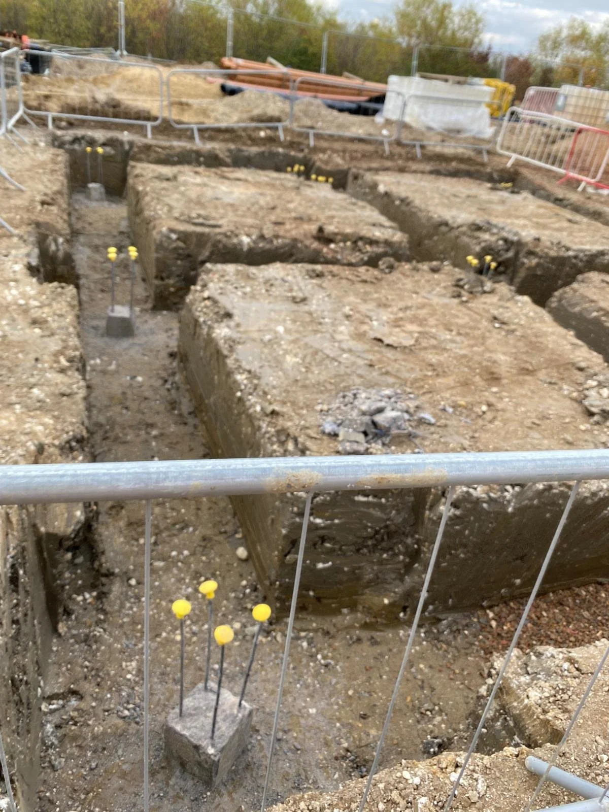 Construction excavation site with trenches and concrete foundations, surrounded by temporary safety barriers and construction materials.