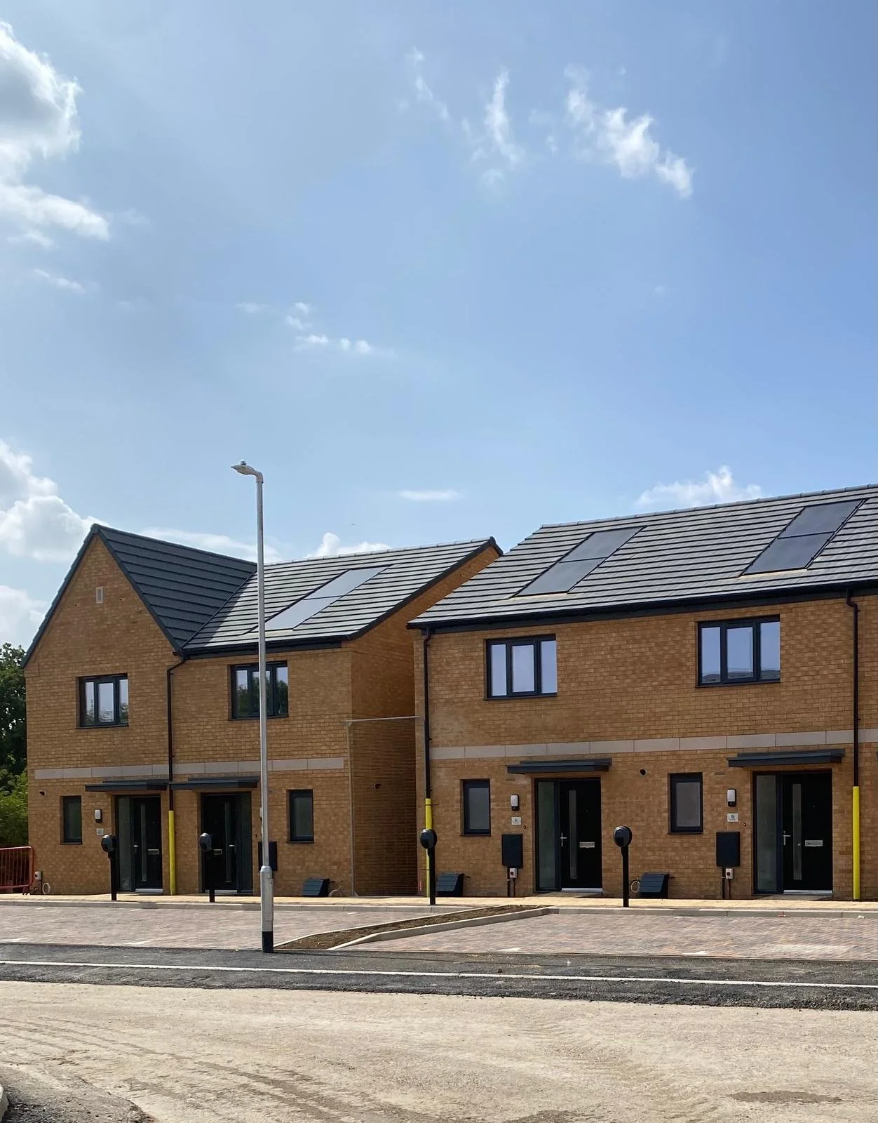 Modern brick houses with solar panels on the roofs, a paved sidewalk, streetlights, and a partially unpaved road under a partly cloudy sky.