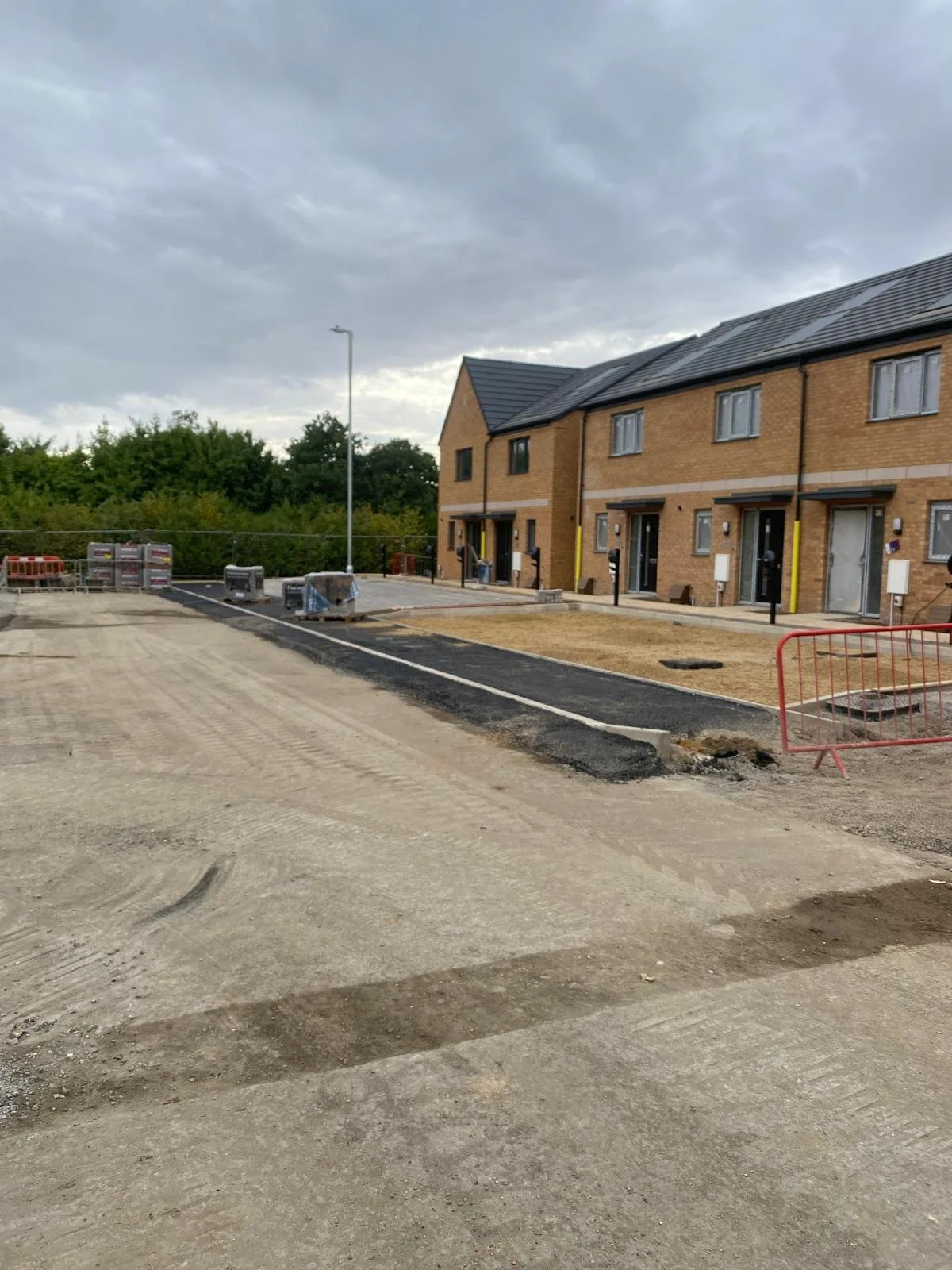 Construction site in front of new brick row houses, with paving being laid and construction materials on site under a cloudy sky.