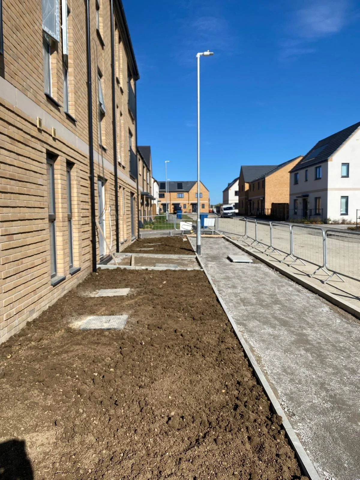 Sidewalk and flower beds under construction with dirt, fencing, and streetlights in a residential neighborhood.