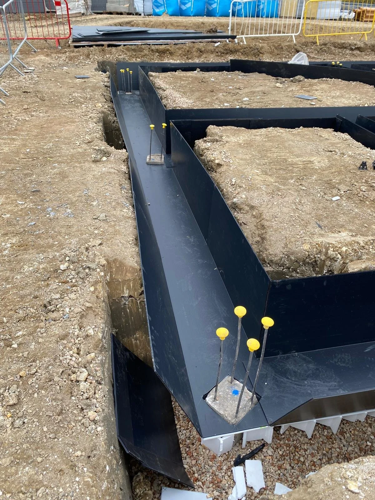Construction site with a black plastic formwork for a building foundation, steel pins with yellow caps securing the form, and excavated dirt around the site.