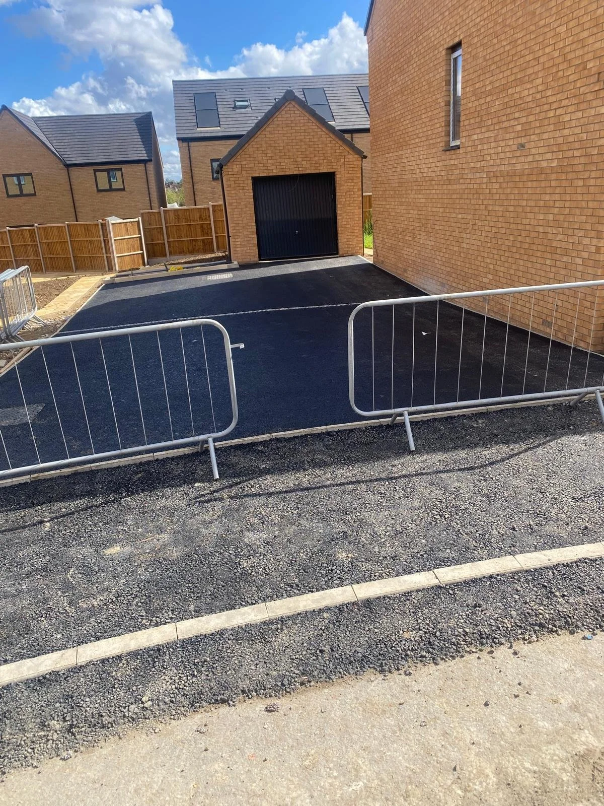 Newly paved driveway in a residential area with wooden fences, brick houses, and a small garage in the background under a partly cloudy sky.